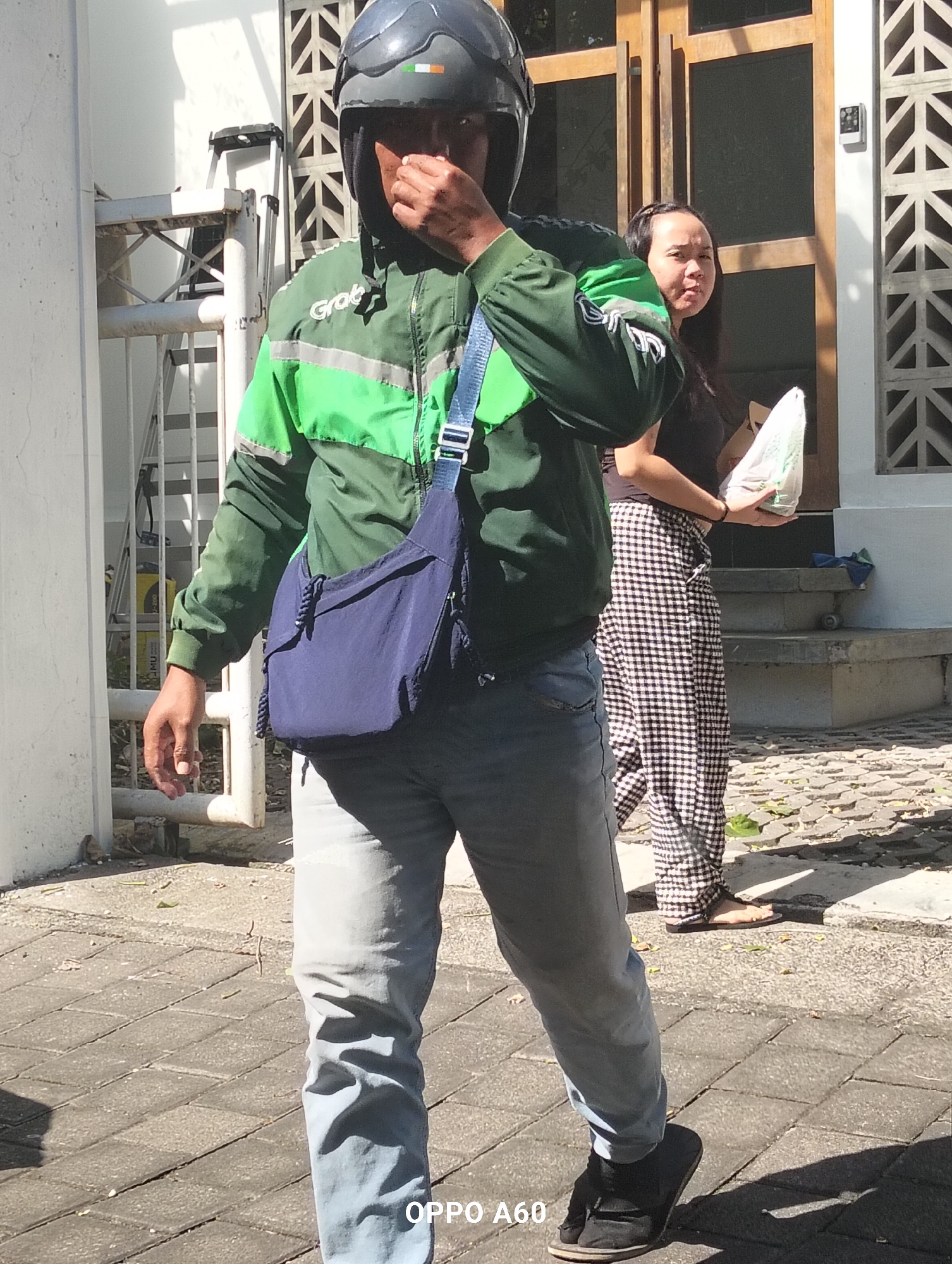 A full shot captures a Grab delivery driver walking on a paved walkway in front of a building in North Kuta, Indonesia. The driver, wearing a black helmet with green and white accents, a dark green Grab jacket with reflective stripes, and light blue jeans, carries a dark blue messenger bag over their left shoulder. The driver's right hand is raised, touching their face. Behind the driver, a woman wearing a dark top and a black and white checkered skirt walks away from the camera, holding a white plastic bag. The environment includes a white wall on the left, a wooden door and a decorative concrete wall on the right, and a set of concrete steps leading to the building. The ground is paved with interlocking stone tiles.