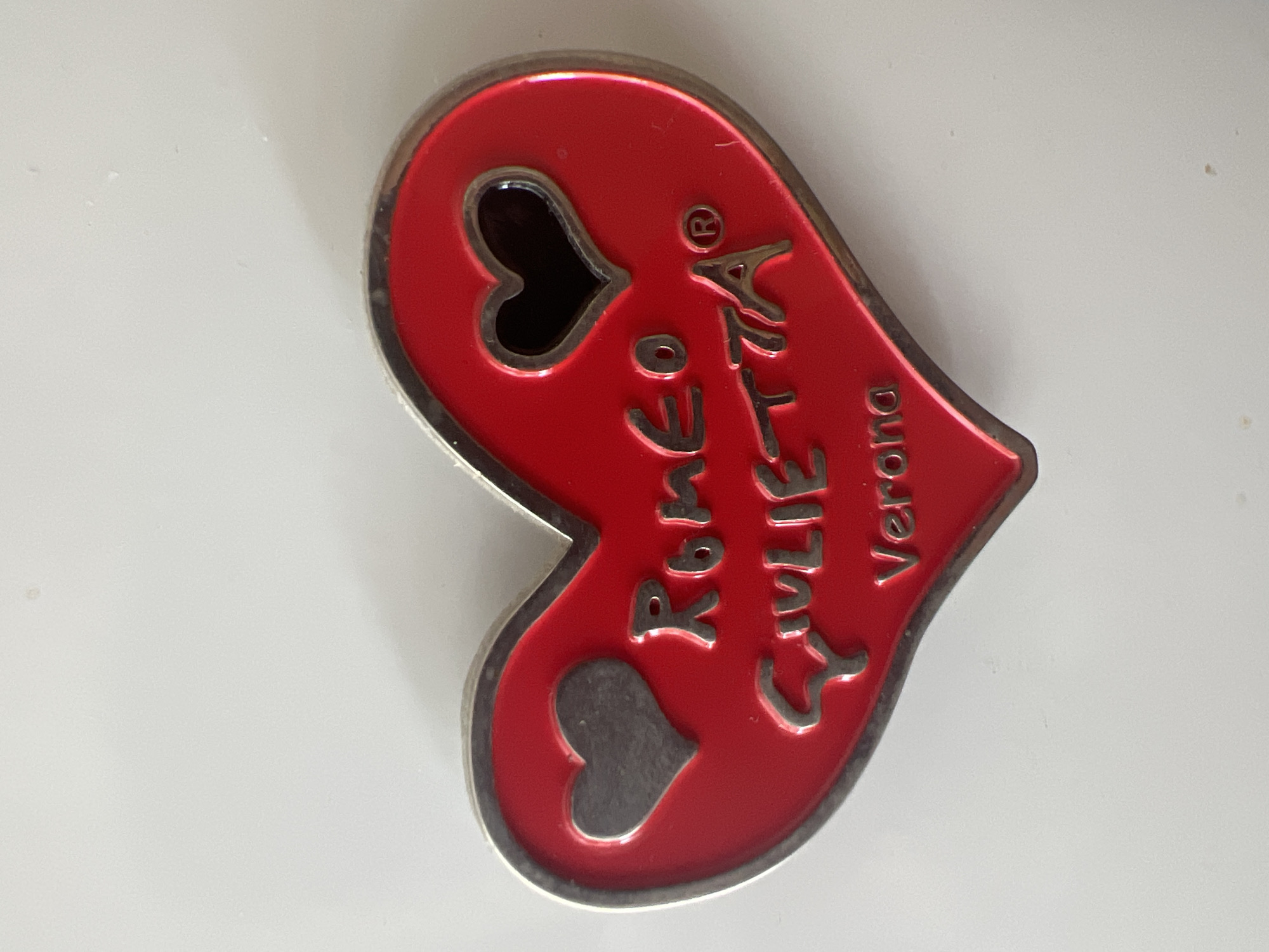 A red heart-shaped pendant with a silver-toned border and a smaller, gray heart shape on the left side of the pendant. In the center of the pendant, the words "ROMEO GIULIETTA" are embossed in silver-toned lettering, followed by a registered trademark symbol. Below "GIULIETTA," the word "Verona" is also embossed in silver-toned lettering. A small, dark heart shape is cut out of the upper right portion of the red pendant. The pendant is lying on a white surface.