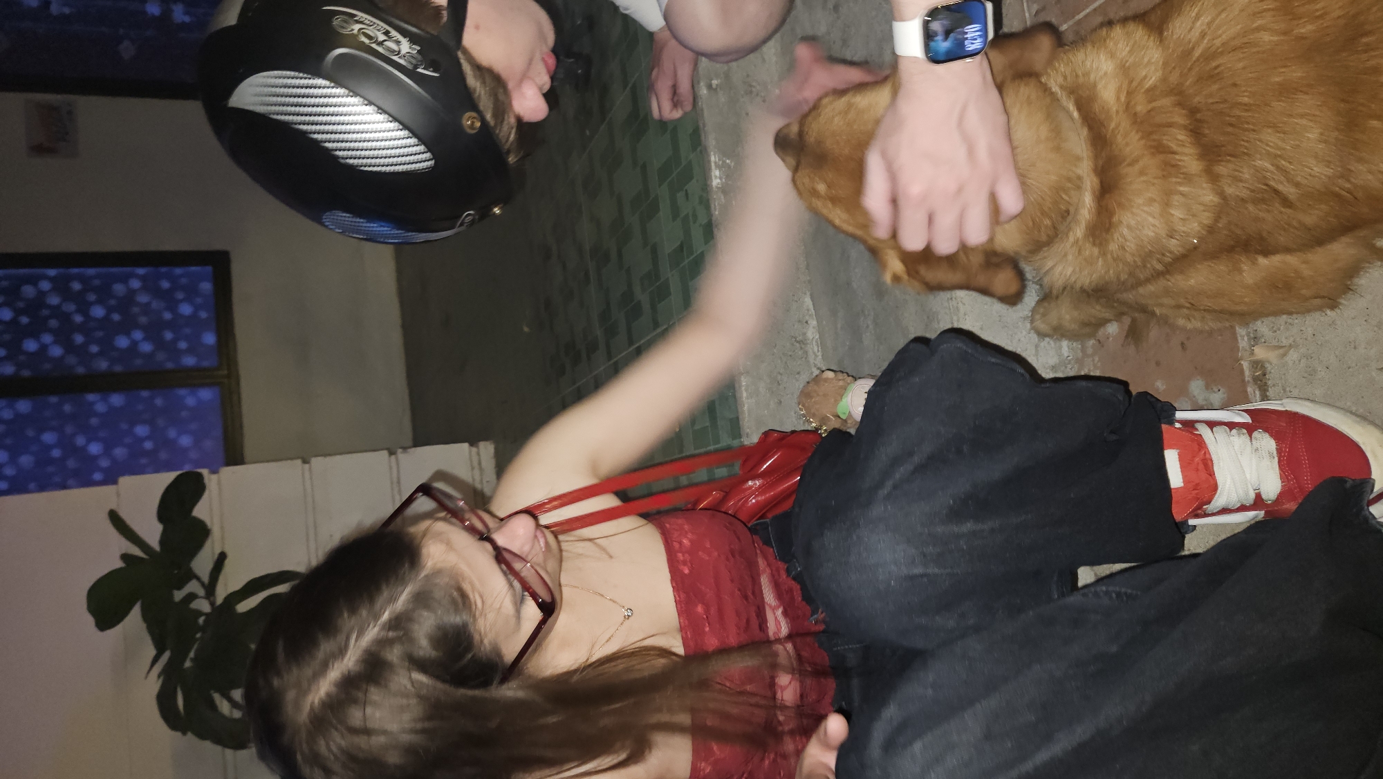 A rotated, wide-angle shot shows several individuals and a dog in what appears to be an outdoor, urban setting.
A young woman wearing glasses and a red lace top is seated on the ground, possibly cross-legged. Next to her is a red bag and a leg covered with dark jeans and red sneakers.
Beside her, an individual in a black helmet is bent over, and next to them, another person wearing a white smartwatch is petting a golden-colored dog. The background features what appears to be a wall and a window.
Although not visible in the picture, based on the surroundings, this could be in Nha Trang, Vietnam.