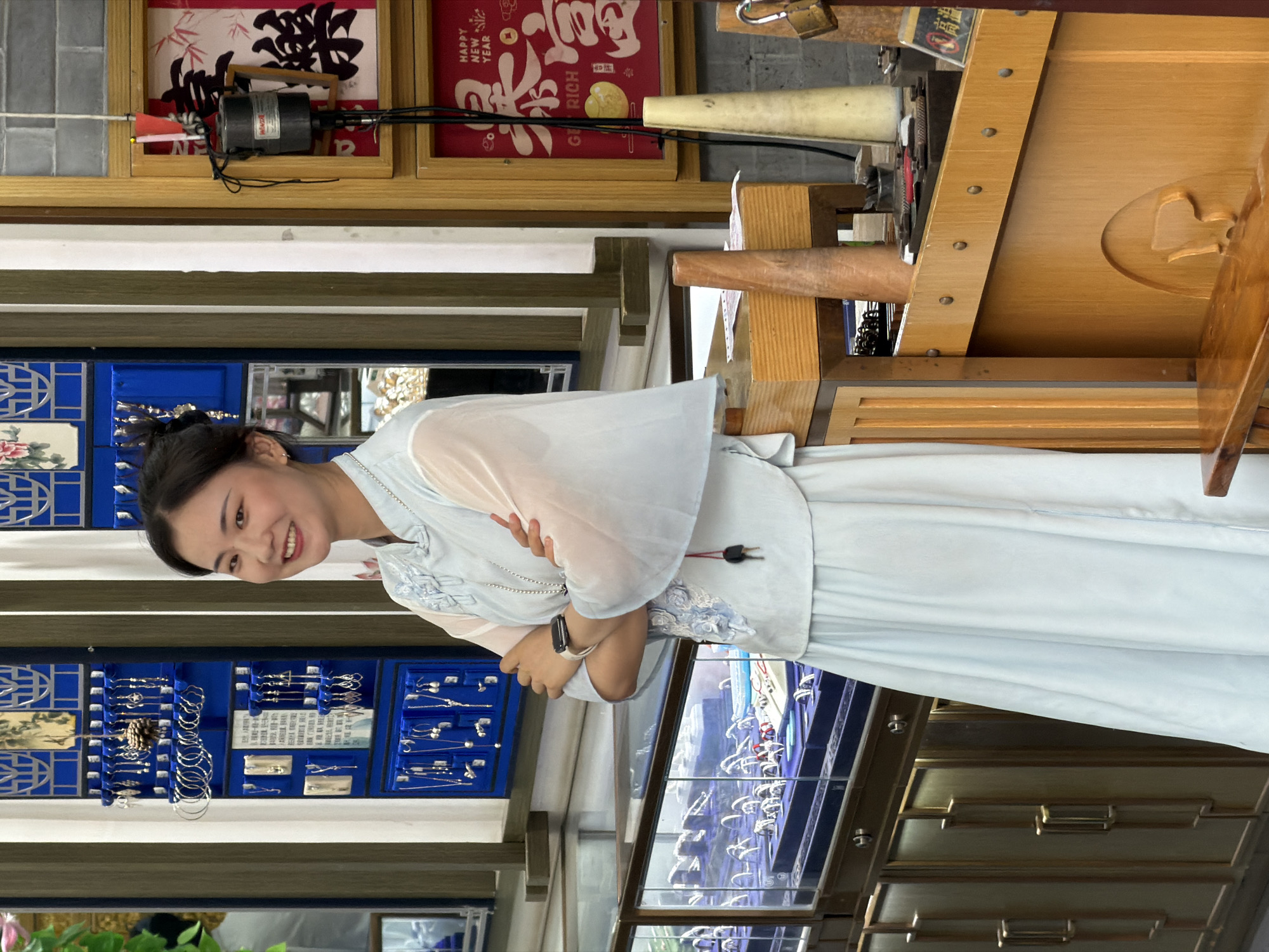 A young woman with dark hair pulled back and a warm smile stands in a shop filled with jewelry. She is wearing a light blue traditional Chinese outfit with sheer sleeves and a long skirt. Her arms are crossed, and she wears a watch on her left wrist. Behind her, a blue display case showcases various silver jewelry pieces, including earrings, necklaces, and bracelets. To her right, a wooden counter holds tools and decorative items, including two conical objects and a framed red sign with Chinese characters that reads "HAPPY NEW YEAR." The shop appears to be a jewelry store in Yuzhong District, China, suggesting a cultural or commercial setting.