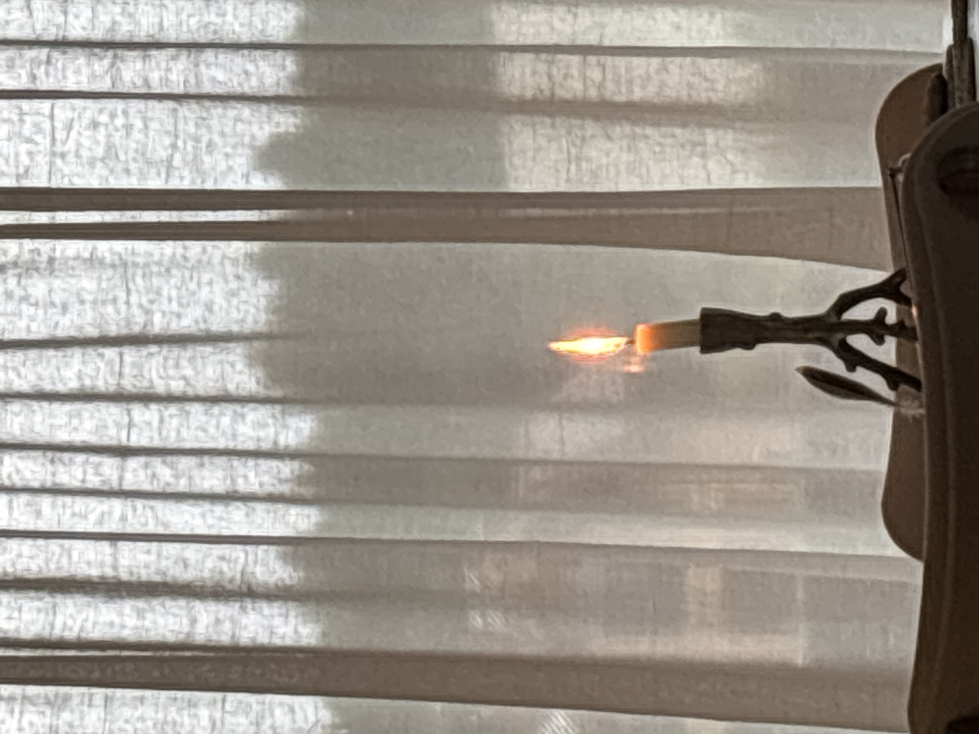 The image depicts a lit candle in a dark, possibly antique, candelabra. The candelabra has a rustic, branch-like design and is made of dark metal. The candle flame is a warm orange and yellow. Behind the candle, sheer white curtains are drawn, with light filtering through them, creating a soft, diffused background. The curtains have vertical pleats. In the foreground, the edge of a dark wooden object, possibly a table or furniture, is visible. A feather quill rests on this object, near the candelabra. The scene appears to be indoors, with the curtains suggesting a window. No specific location in Zvenigorod, Russia, can be identified from this image.