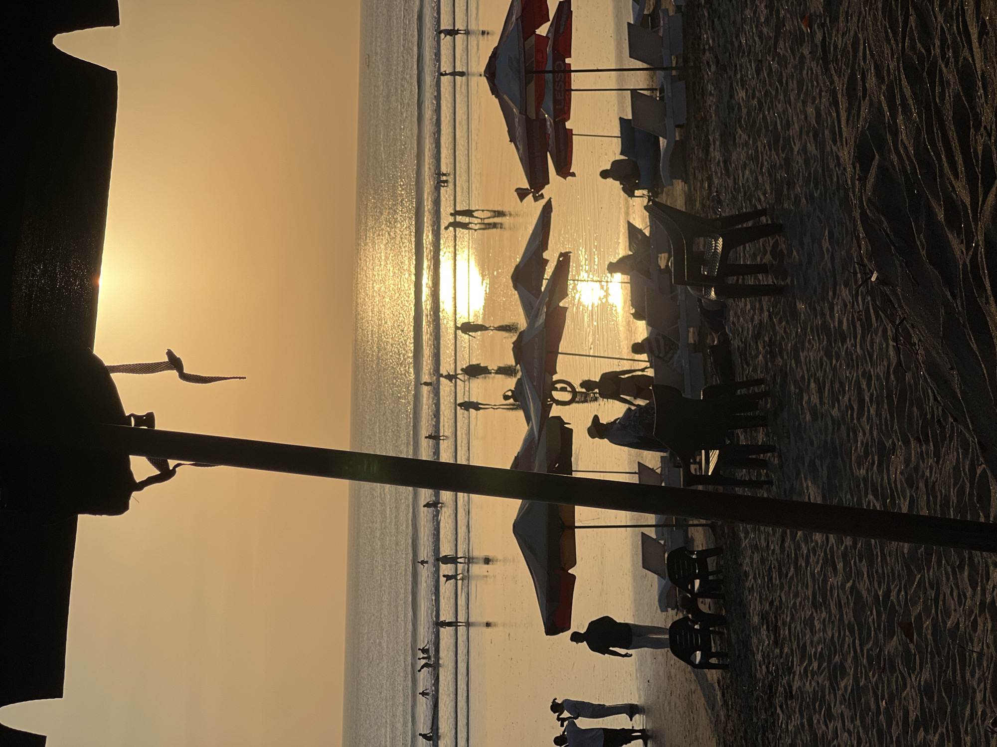 This is a horizontal, medium shot, taken at sunset on Kuta Beach, Indonesia, as indicated by the golden hour light reflecting off the ocean and the presence of beachgoers. The foreground is dominated by dark silhouettes of beach umbrellas and chairs on the sandy shore. In the midground, several people are walking along the wet sand, their reflections shimmering in the water. Further out, multiple individuals are wading and playing in the gentle waves. The ocean stretches to the horizon, with a bright sun setting, casting a warm, orange glow across the sky and water. The atmosphere is tranquil and evokes a sense of leisure and end-of-day relaxation.