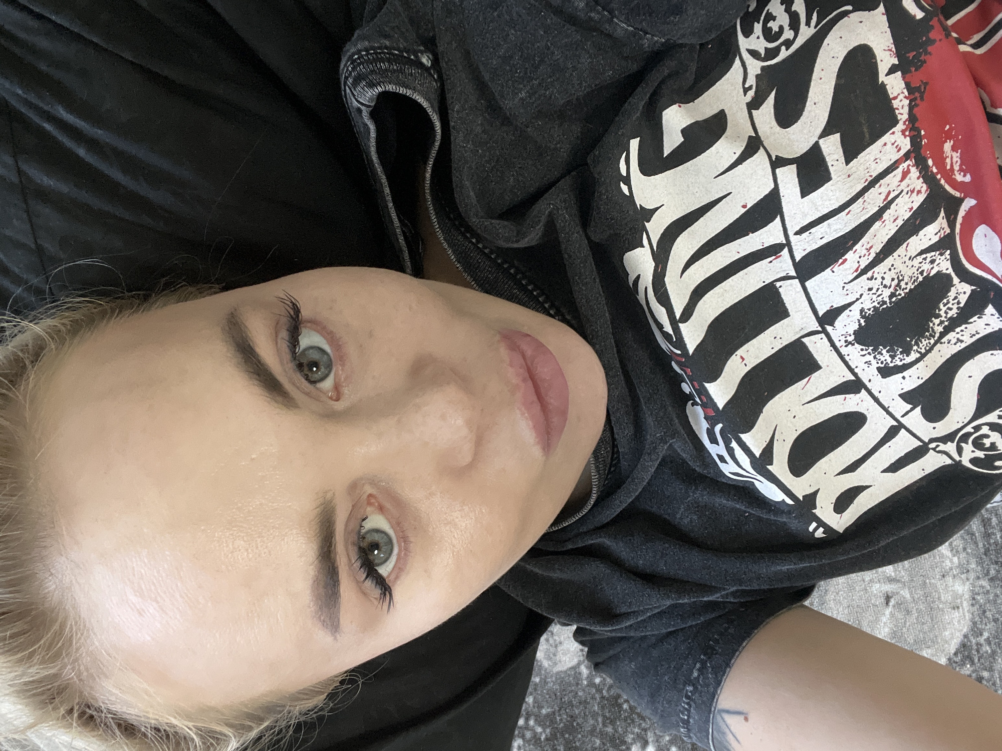 An overhead, close-up selfie is captured from a high angle looking down on a young woman lying down. The woman has blonde hair pulled back from her face. She is wearing a dark gray t-shirt with a Rolling Stones logo on it. Her head is tilted slightly, and she is looking towards the camera with her eyes open. Her lips are slightly parted, and she has on light makeup, including mascara and lip gloss. The background appears to be a dark, textured surface, possibly a couch or blanket, and a floor with a gray and white pattern. The lighting is bright, illuminating her face and clothing. There are no visible signs of the city or any specific location.