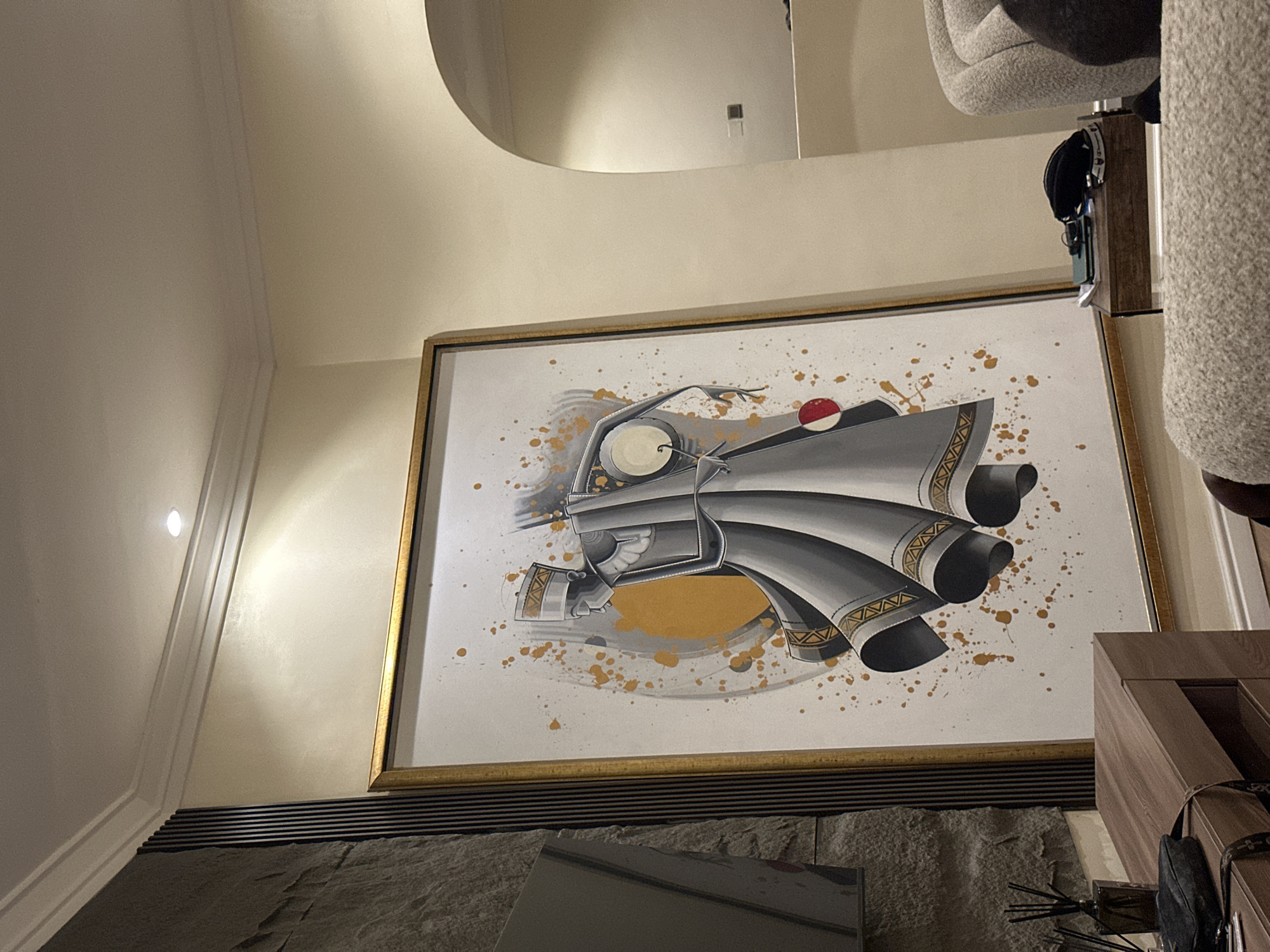A framed painting hangs on a wall above a dark gray surface. The painting depicts a stylized, gray and white drawing with gold accents. The wall is painted a pale yellow. A light fixture is visible on the left wall. Part of a chair is visible in the upper right corner. The scene's location is unknown and there is no evidence it is in Ilado, Nigeria.