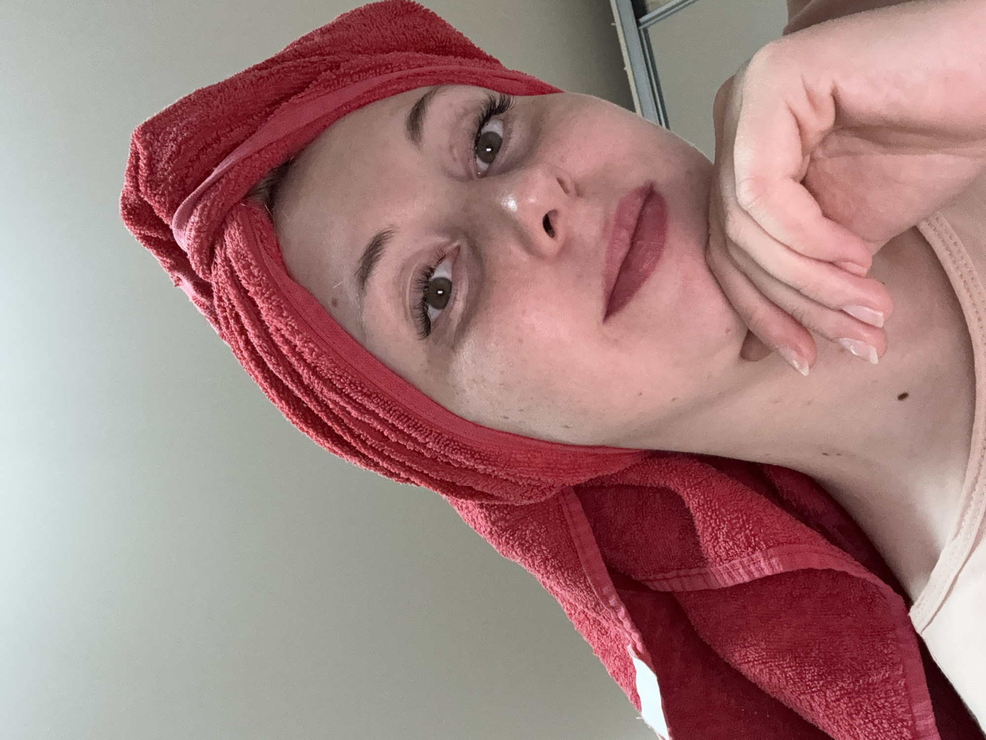 A low-angle close-up photograph shows a person with a red towel wrapped around their head. The person's face is visible, with their left hand positioned near their chin. The background appears to be a plain, light-colored surface. The person is indoors.