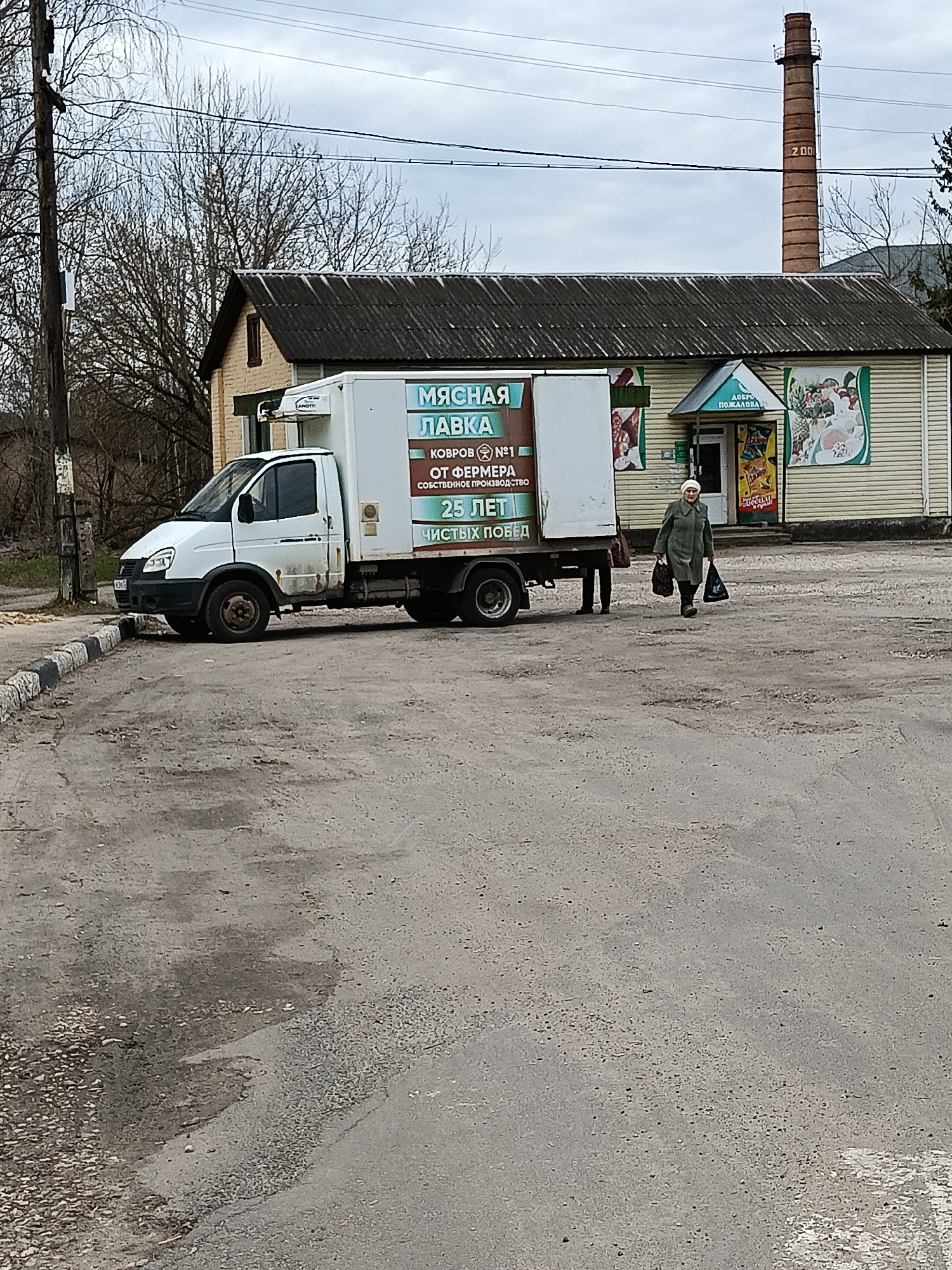 The image depicts a street scene in Красный Маяк, Russia. A white, box-truck parked next to a building, the side of which is painted with advertisements for a "Meat Shop" ("МЯСНАЯ ЛАВКА"), "Rugs #1" ("КОВРОВ №1"), and "25 years of clean victories" ("25 ЛЕТ ЧИСТЫХ ПОБЕД"). A woman carrying two plastic shopping bags is walking in front of the truck. The building next to the truck has a green awning above the entrance that reads "Welcome" ("ДОБРО ПОЖАЛОВАТ"). To the right of the entrance are posters with images of food. Behind the building, there is a brick smokestack with the number "200" written on it. The sky is overcast. The street is paved with asphalt.
