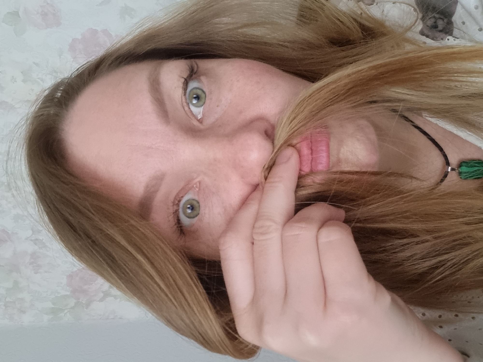 A light-skinned woman with blonde hair is photographed from an upside-down perspective. Her green eyes are prominent. She is holding a strand of her hair in front of her mouth, distorting her lips. She wears a black cord necklace with a green pendant. A floral-patterned fabric is visible in the background. The location is Chaikovsky, Russia.