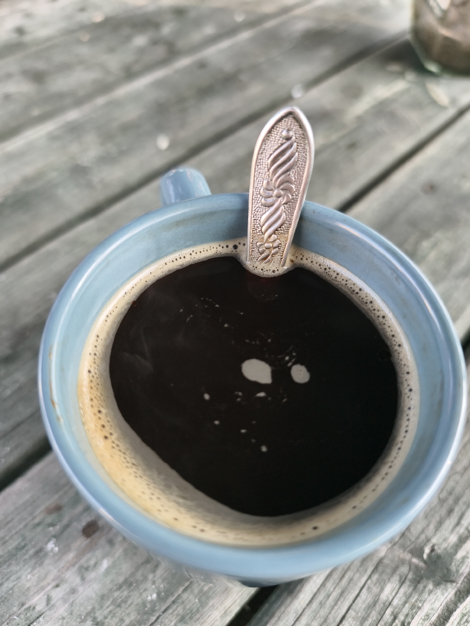 An overhead, close-up shot shows a light blue ceramic mug filled with dark coffee, resting on a weathered, light green wooden surface. A silver spoon with an ornate floral design is positioned in the coffee. The coffee has a thin layer of foam around the rim. The wooden surface appears to be part of an outdoor table, possibly in a park or garden. There are no people or animals visible in the image. The lighting suggests it is daytime, with natural light illuminating the scene. The image does not provide any information about the city of Tczew, Poland.