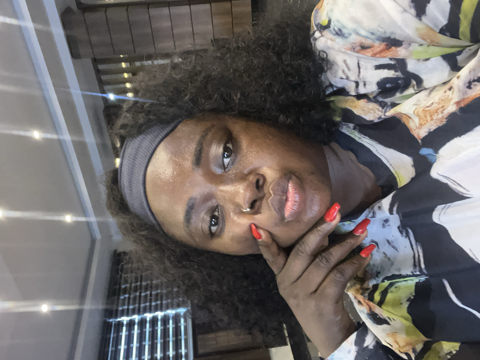A medium shot captures a woman from the chest up, facing the camera at a slightly downward angle. She has dark, curly hair pulled back with a grey headband. Her skin is dark, and she wears bright red nail polish on her long fingernails. One hand is placed on her cheek, with her fingers gently touching her face. She has a subtle nose ring. Her lips are slightly parted, as if she's about to speak. She is wearing a colorful, patterned blouse with shades of black, white, yellow, orange, and blue.
The background appears to be an indoor setting, possibly an office or lobby. To the left, venetian blinds are visible behind a window, suggesting a view to the outside or another room. The ceiling is white with recessed lighting. To the right, vertical wooden panels are part of the interior design. The overall lighting is bright, with some light reflections on the ceiling. This scene does not provide enough information to determine its specific location within Abuja, Nigeria.