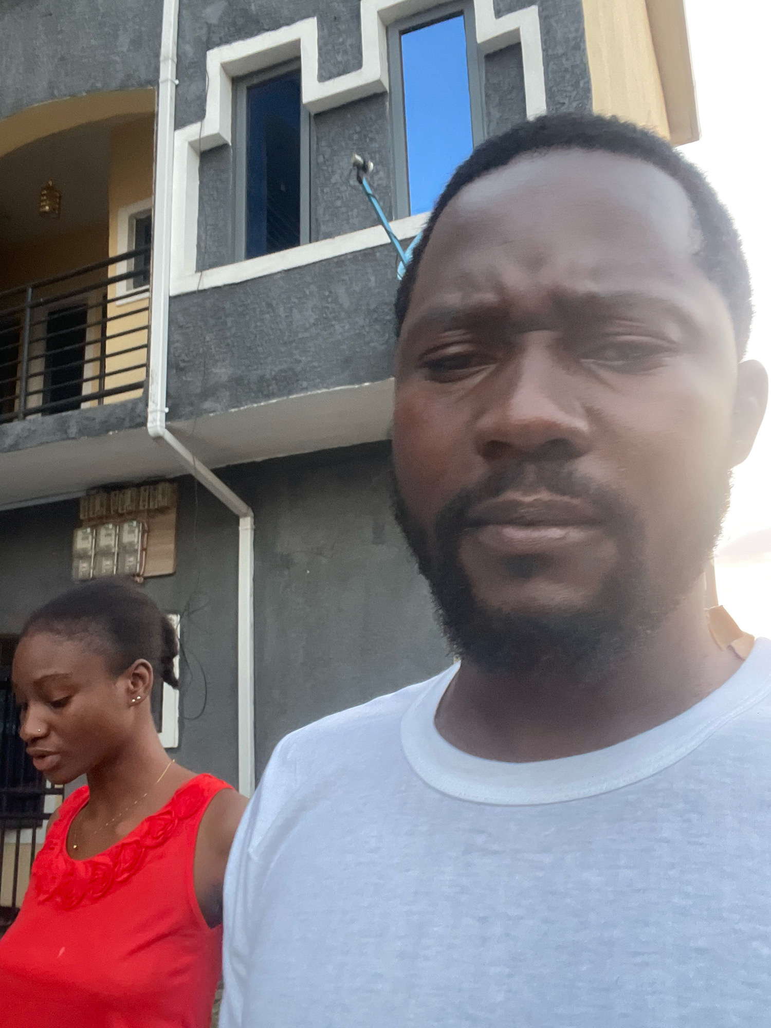 In an outdoor, daytime scene in Abuja, Nigeria, a man with dark skin, a beard, and short dark hair is the primary subject, positioned close to the camera and looking slightly off-center. He is wearing a white t-shirt. To his left and slightly behind him is a young woman with dark skin and dark hair pulled back in a ponytail. She is wearing a bright red sleeveless top with a decorative ruffled neckline and a thin gold necklace. Both individuals appear to be standing in front of a building with grey and beige exterior walls. The building features windows, a balcony with metal railings, and visible electrical conduits and a junction box. The lighting suggests it is likely a sunny day.