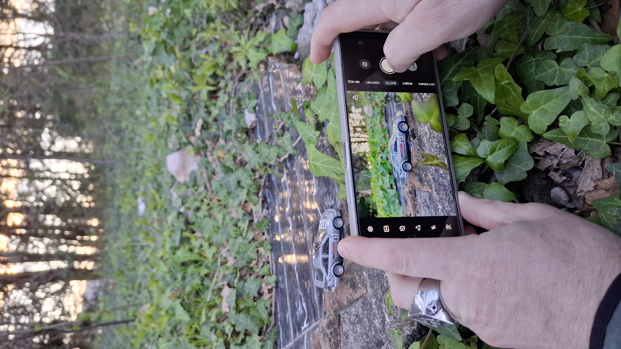 A person is taking a photo with a smartphone of a toy car. The car is silver and gray with the number "87" on its side. It is placed on a stone surface, likely part of a walkway or wall, which appears damp and reflective. The background includes ivy and other green foliage. The person holding the phone has pale skin and is wearing a ring on their finger. The screen of the phone shows a live view of the car and surroundings, indicating the photo is being taken. The environment is outdoors, with a blurry view of trees in the background and lighting suggesting it's either early morning or late afternoon. The location is not identifiable in the city of Unknown, Unknown, based solely on the provided image.