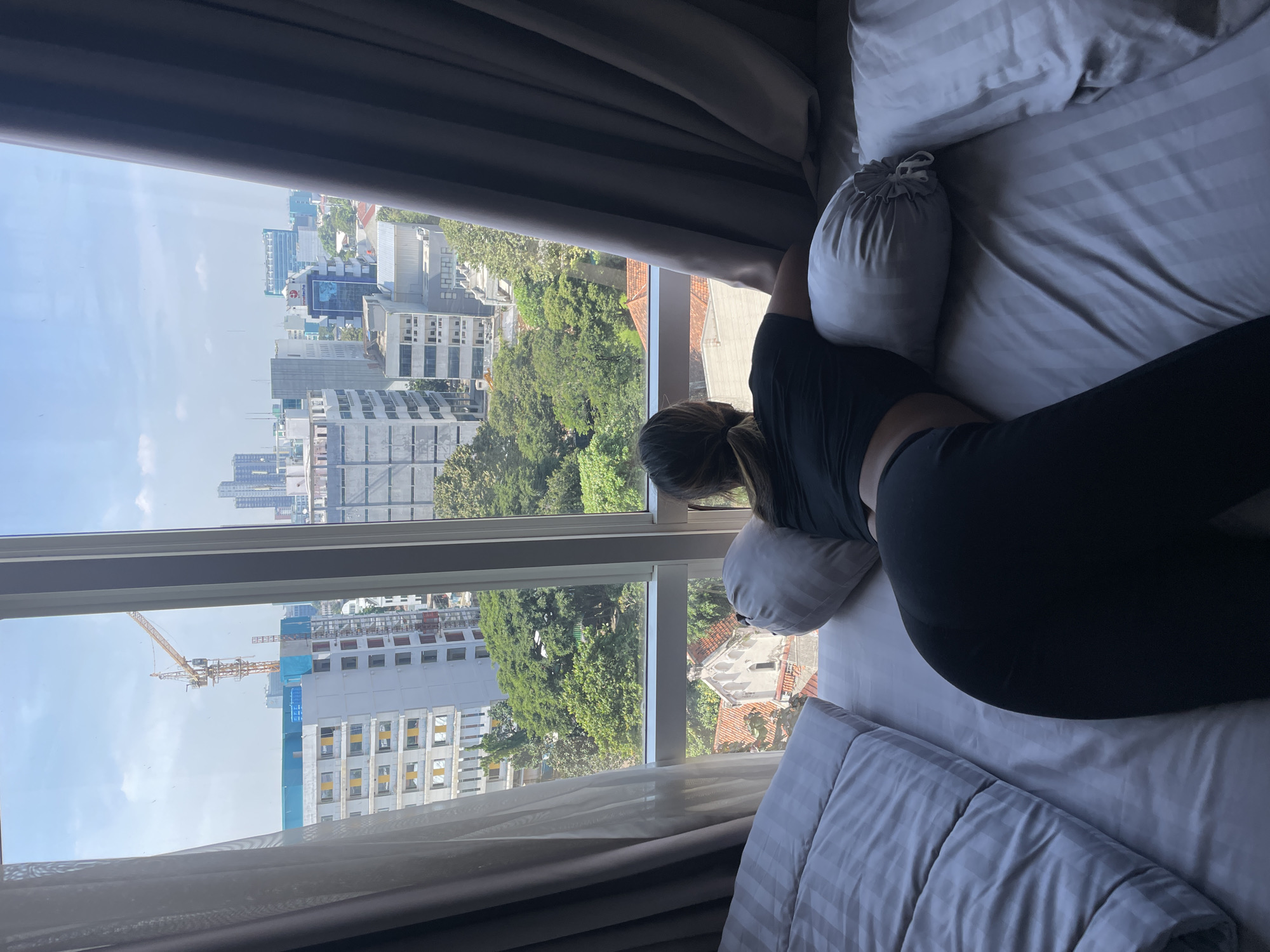 A person is lying on a bed, oriented to the right of the frame, next to a window with a view of a cityscape. The person is wearing a black top and black leggings, with their head near the window. The bed has white striped bedding and several pillows. Outside the window, there are buildings of varying heights, some with visible construction, and green trees. The buildings suggest an urban setting. The location is Central Jakarta, Indonesia.