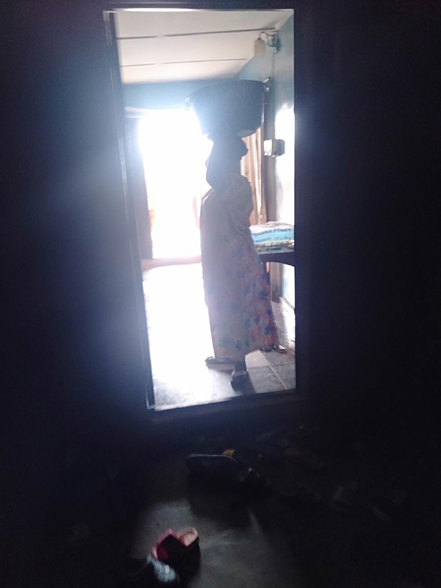 The image shows a person standing in a doorway with a container on their head. The person is wearing a long, patterned dress. A table is visible behind the person, with a blue and white object on it. The room outside the doorway is brightly lit. The room from which the photo is taken is dark, with footwear visible on the floor. The location is Unknown, Unknown.