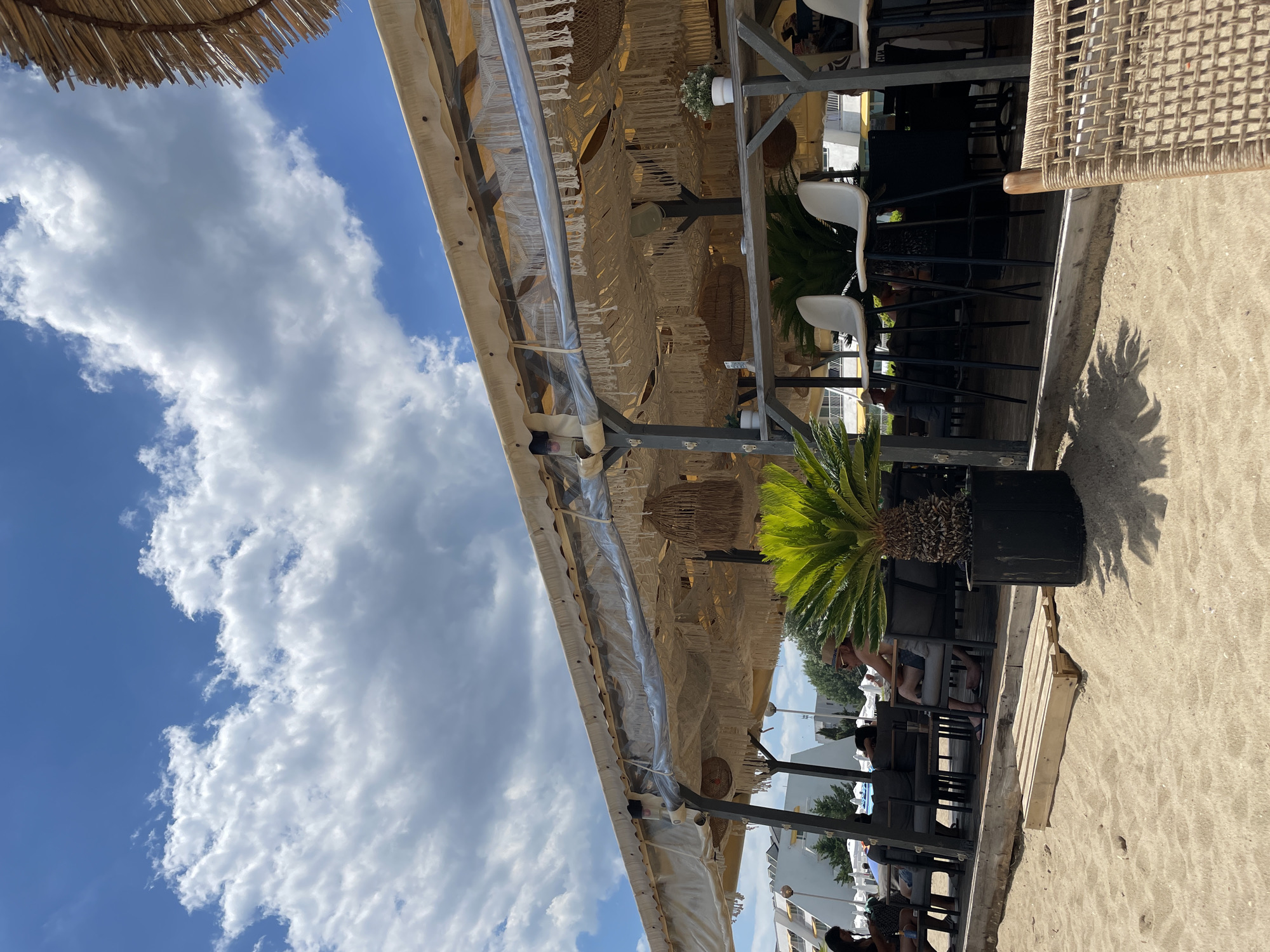 The image shows a beachside cafe in Obzor, Bulgaria. The cafe has a covered seating area with a thatched roof and fabric draping decorated with macrame. White bar stools are positioned at a counter, and dark seating is arranged on a wooden platform behind it. A potted palm tree stands in front of the cafe. In the background, people are seated at tables, and some buildings are visible. The ground is covered in sand, and the sky is blue with fluffy white clouds.