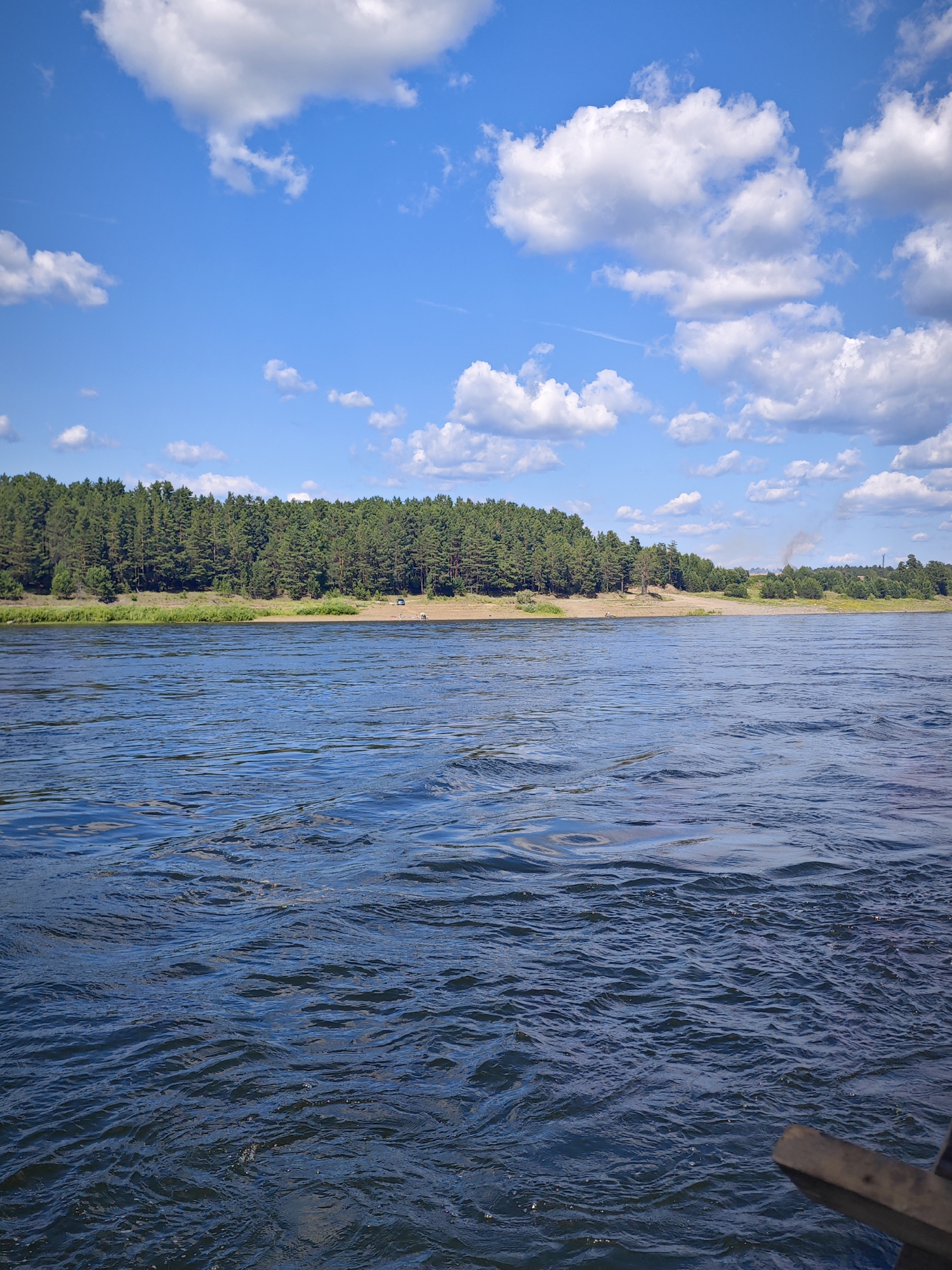 The image captures a serene river landscape under a bright blue sky dotted with fluffy white clouds. The foreground is dominated by the dark blue, slightly rippled water of a wide river. In the middle ground, a sandy shore is visible, backed by a dense forest of green pine trees that stretches across the horizon. A few small figures, possibly people, are present on the shore, along with a dark-colored vehicle and what appears to be a light-colored object. In the far distance, a faint plume of smoke rises from behind the trees, suggesting some distant industrial activity. The overall scene depicts a tranquil natural environment, likely in Abalakovo, Russia, given the context.