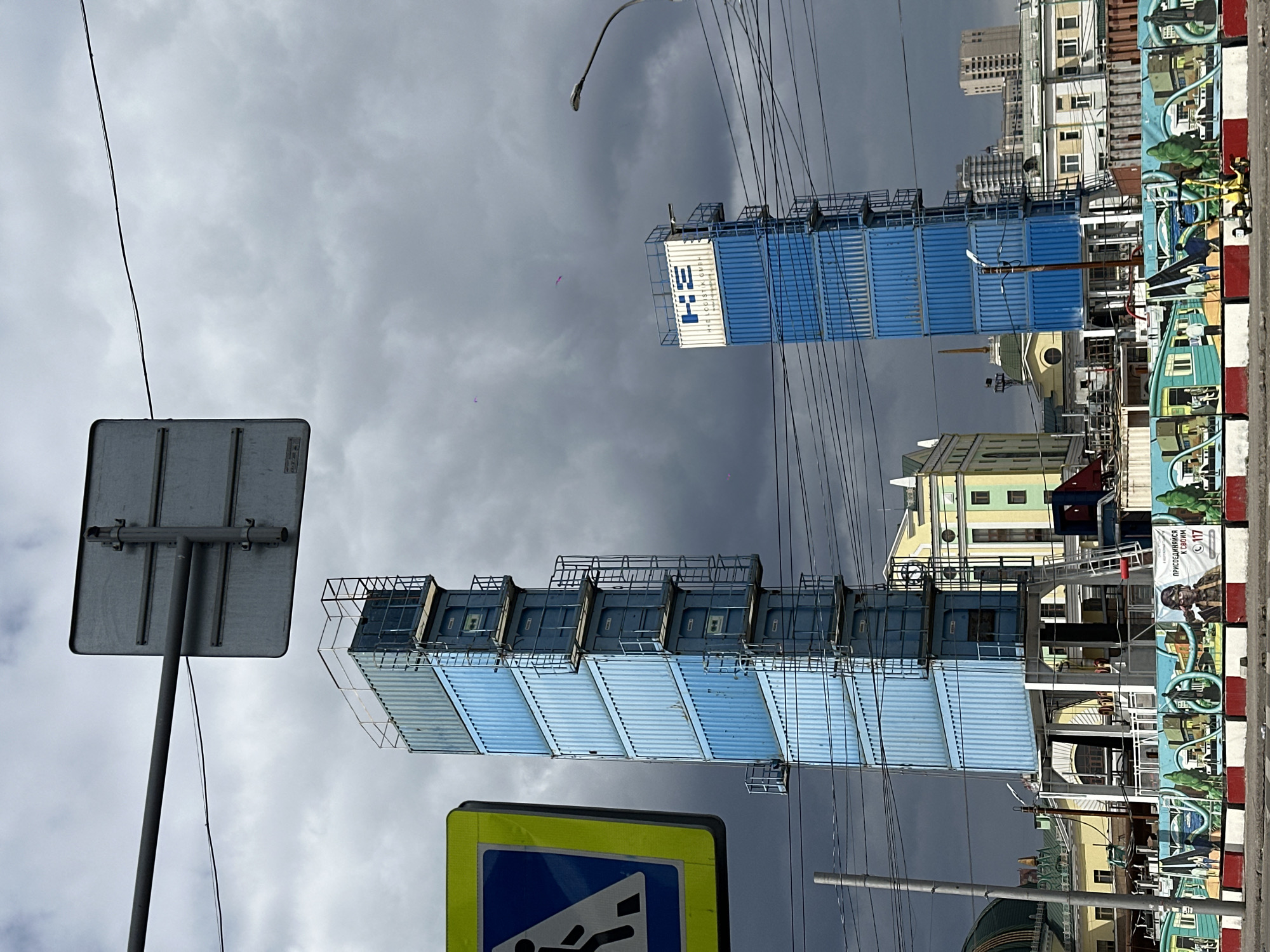 Here is a description of the image.
The image shows a view in Krasnoyarsk, Russia. Two structures made of stacked blue shipping containers are visible. These structures appear to be placed near a street with a pedestrian crossing sign. The sky is overcast. Buildings with varying architectural styles are behind the structures. A decorated concrete barrier with cartoonish murals is also visible to the right of the scene.
