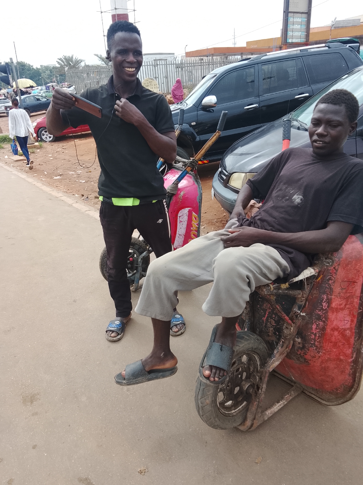 Two African men are standing next to an improvised tricycle on a city street. The man on the left is smiling and holding a cell phone. He is wearing a black shirt, black pants with a neon green stripe at the waist, and sandals. The man on the right is seated on the tricycle, wearing a dark shirt and light-colored pants, also wearing sandals. The tricycle is red and appears to be made from a wheelbarrow. In the background, there are parked cars, a building with signage, and a fenced area with pedestrians. The location is identified as the city of Abuja, Nigeria.