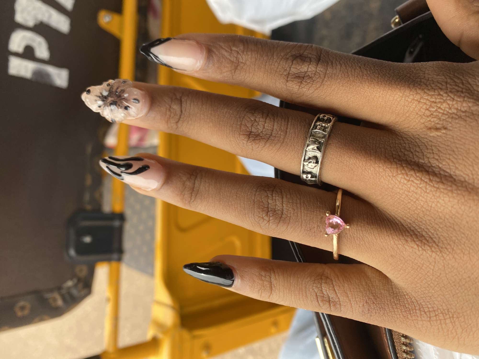 The image shows a close-up view of a person's hand with manicured nails. The nails are long and feature various designs: one has a white and black floral embellishment, another has a black and white tribal pattern, some have a French tip with black accents, and one is solid black. Two rings are visible on the fingers: a silver band with embossed designs and a delicate gold ring with a small pink heart-shaped stone.
The hand is positioned in front of a blurred background that appears to be a bright yellow object, possibly a vehicle or street furniture, and a dark surface with some lighter patterns. The lighting suggests it is daytime, and the overall impression is casual. The focus is on the intricate nail art and the jewelry. No specific activity or event is clearly depicted beyond the presentation of the hand and its adornments. There is no visible text in the image. The context provided suggests the image was taken in Jos, Nigeria.