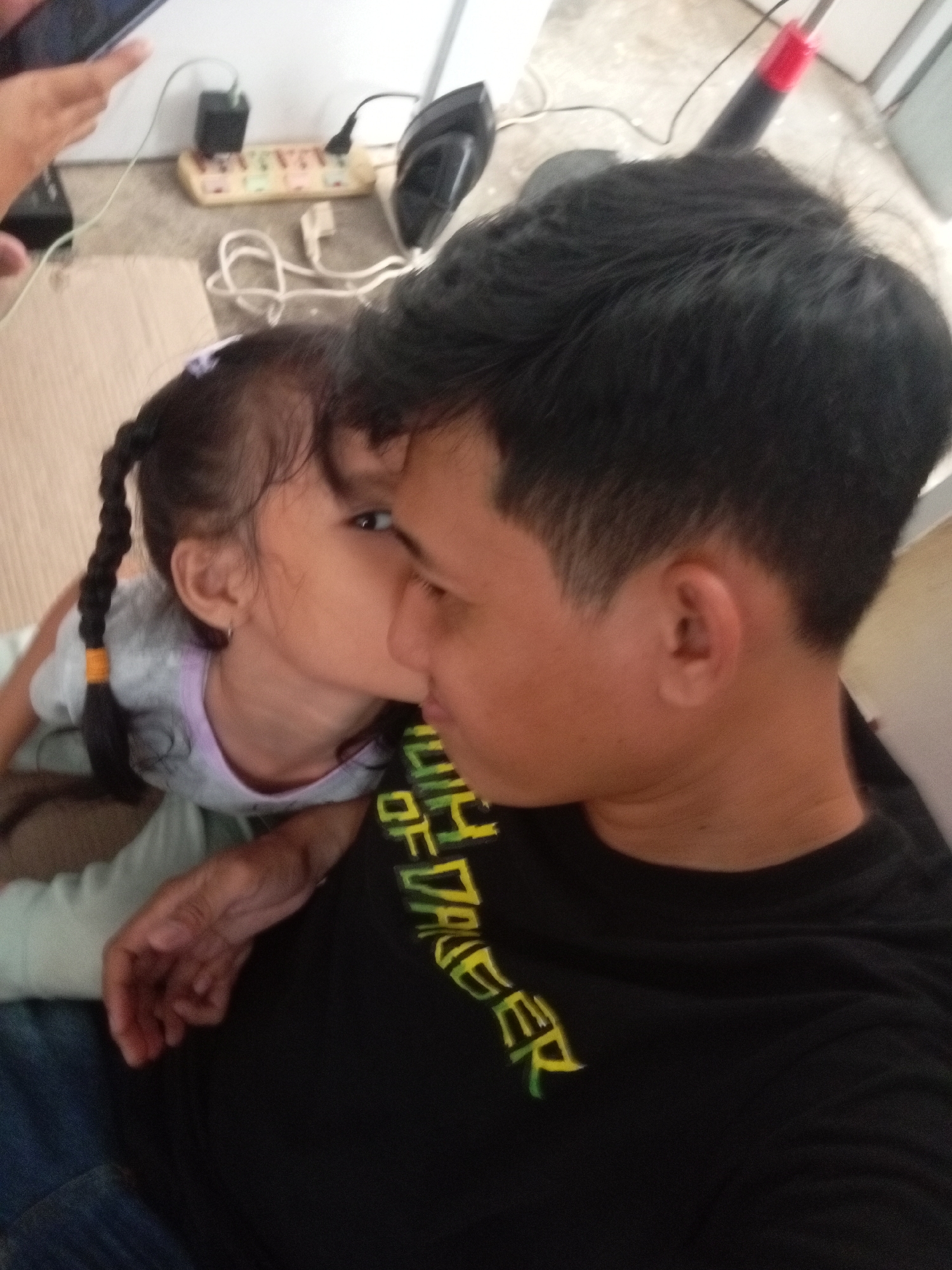 The image shows a close-up of a young child leaning in to kiss the cheek of a young man. The child has dark hair in a braid and is wearing a light-colored shirt with a purple trim. The man has dark hair and is wearing a black t-shirt with green and yellow text on it. The text appears to read "DAREDEVIL" or something similar.
The background shows a floor with a power strip plugged into an outlet, and a black cord extending from it. There are also hints of white and gray objects, possibly furniture. The lighting suggests it's daytime, but no specific time is indicated. The overall mood appears to be affectionate and familial. The scene is likely indoors, possibly a home. The location context is Batam, Indonesia.