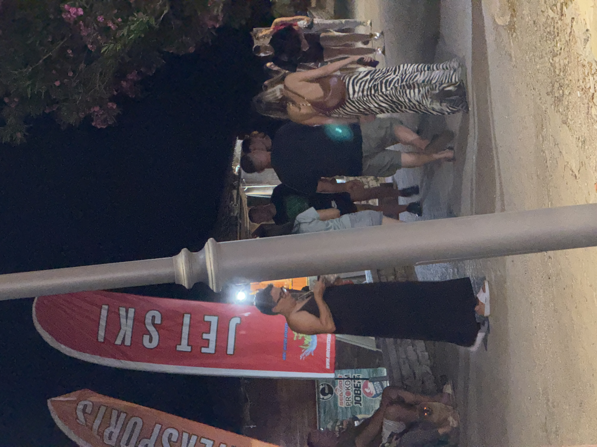 This media file shows a group of people standing and walking on a paved walkway at night, illuminated by artificial lighting. The scene appears to be a lively outdoor area, possibly a promenade or street in a tourist destination.
In the foreground, a woman in a black dress and glasses is holding a phone, looking towards her right. Next to her, a red banner with white text reads "JET SKI". Below that, an orange banner with white text is partially visible, showing "ORTS". A large, silver-colored metal pole runs horizontally across the middle of the image.
Further back, several other individuals are visible. One person is wearing a zebra-print dress and another is in a dark shirt and shorts. They appear to be socializing or moving along the walkway. The background is dark, suggesting it is nighttime. The ground appears to be a rough, light-colored pavement. The presence of "JET SKI" and "SPORTS" on banners suggests a location associated with water sports or recreational activities. The overall atmosphere seems casual and possibly festive, typical of a summer evening in a resort town.