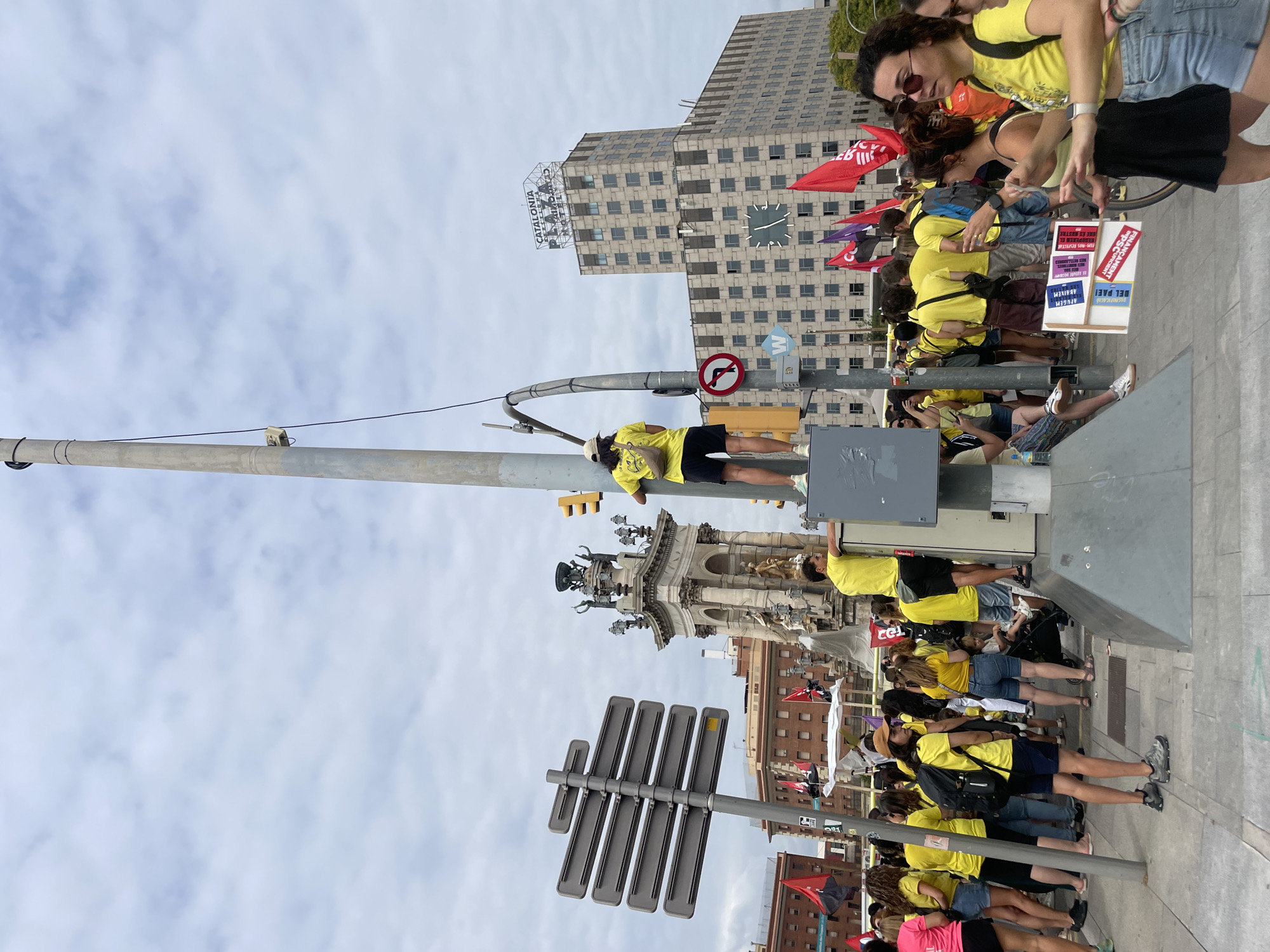 The image depicts a public gathering in Barcelona, Spain, likely in or near Plaça de Catalunya, given the presence of the Catalunya Plaza building visible in the background. A large crowd of people, many wearing yellow shirts, are present on a paved area. A prominent central feature is a tall lamppost with traffic lights and various signs attached to it. A person in a yellow shirt and black shorts stands on top of a gray utility box attached to the lamppost, facing away from the camera. Another person in a yellow shirt and dark shorts is on the lamppost's base.
In the background, a large, ornate monument with statues and columns stands prominently. Behind this monument and the crowd, the "Catalunya Plaza" building, characterized by its grid-like facade and a rooftop sign, dominates the skyline. Red and purple flags are visible, some held aloft by individuals in the crowd and others attached to poles.
On the ground in the foreground, several people are walking and interacting. One person is holding a sign with text in Catalan. A bicycle is partially visible. The sky is overcast with clouds. The overall scene suggests an organized event or demonstration.