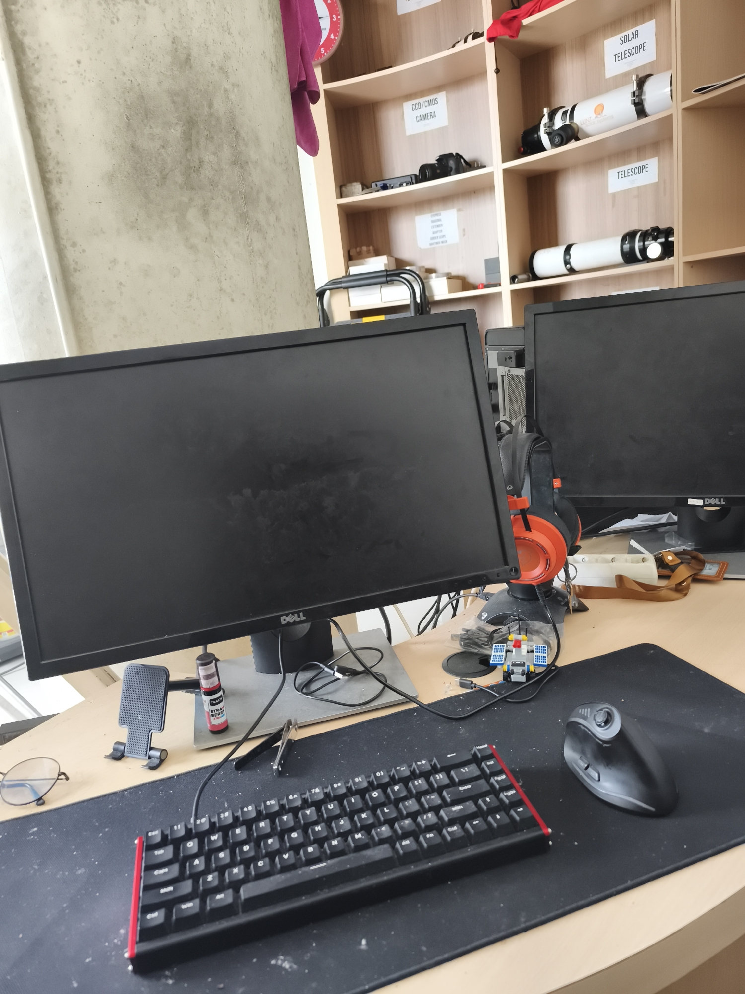 A desk with two computer monitors is pictured. The monitor on the left is a Dell brand. In front of the left monitor is a black keyboard with red accents. A black mouse sits on a black mousepad to the right of the keyboard. On the desk are also a pair of glasses, a small Lego-like toy, a phone stand, and a small bottle. Behind the monitors are shelves filled with various items, including what appear to be telescopes and cameras. The background wall is textured and off-white. No people are visible in the image. The lighting suggests it is daytime, with natural light entering from the left. The environment appears to be an indoor workspace or hobby room.
