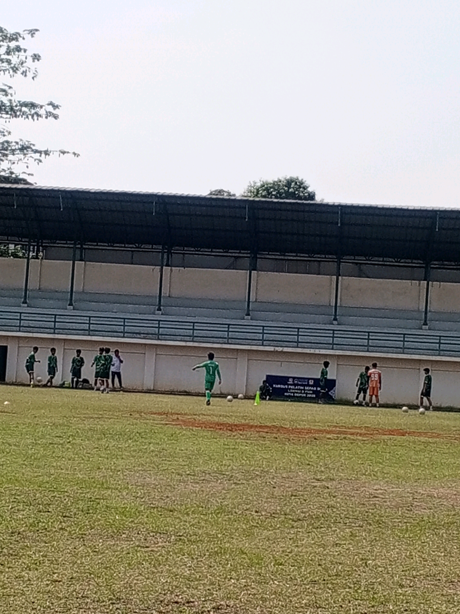 A group of young boys in soccer uniforms are practicing on a grassy field in front of a stadium with bleachers. The boys are in various positions, some running, some standing, and some appear to be kicking a soccer ball. The field is marked with orange lines. In the background, there is a banner that reads "KURUS PELATIHAN SEPAK BOLA" and "KOTA DEPOK 2023". The sky is bright and overcast.