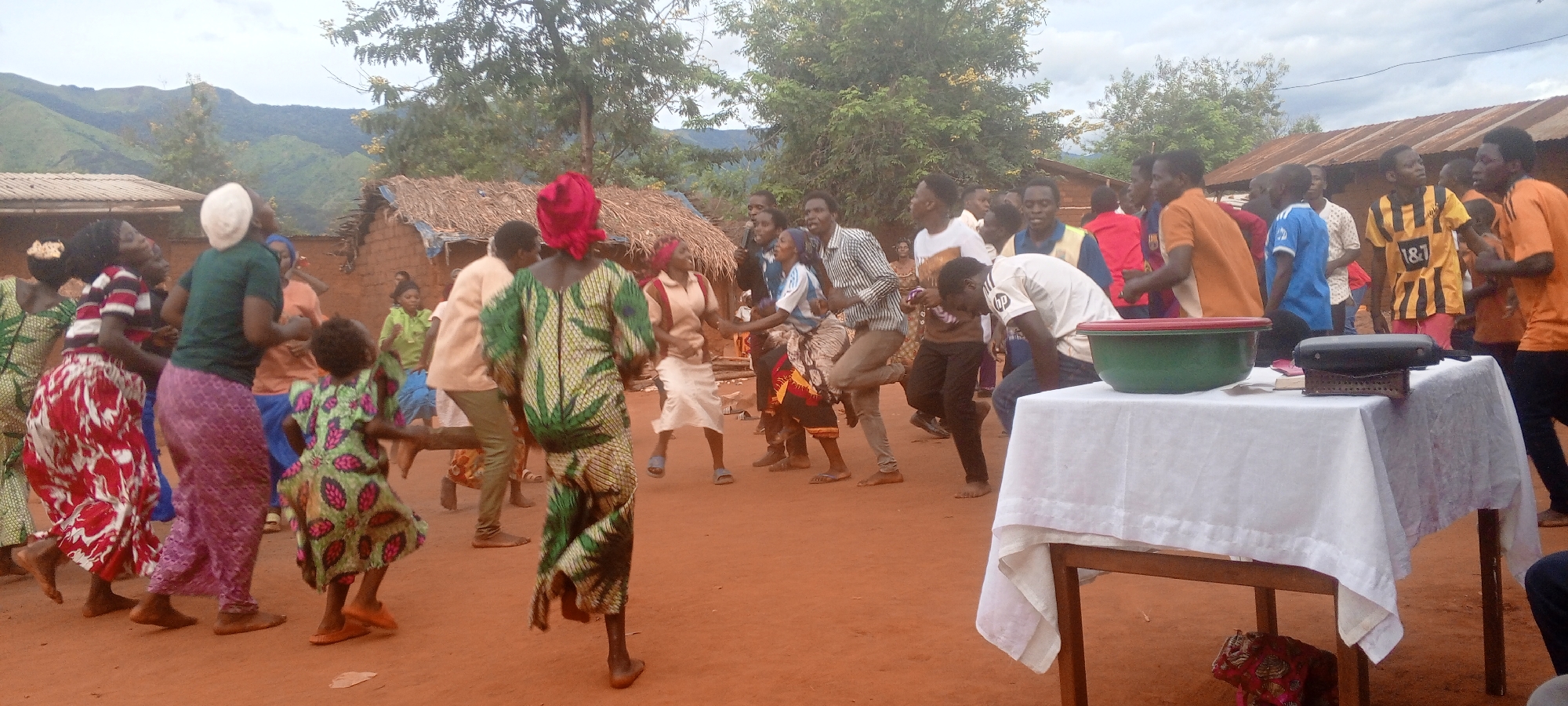 In Ngalula, Democratic Republic of the Congo, a gathering of people is taking place in an outdoor setting. The group is composed of both men and women of varying ages, some of whom appear to be engaged in dancing. The women are wearing colorful, patterned dresses or skirts, while the men are dressed in a mix of casual attire, including shirts with sports team logos.
In the foreground, there is a table covered with a white cloth. A green bowl and a black object are placed on top of the table. In the background, there are trees and buildings with thatched roofs and a mountain range is visible in the distance. The ground is a reddish-brown color.