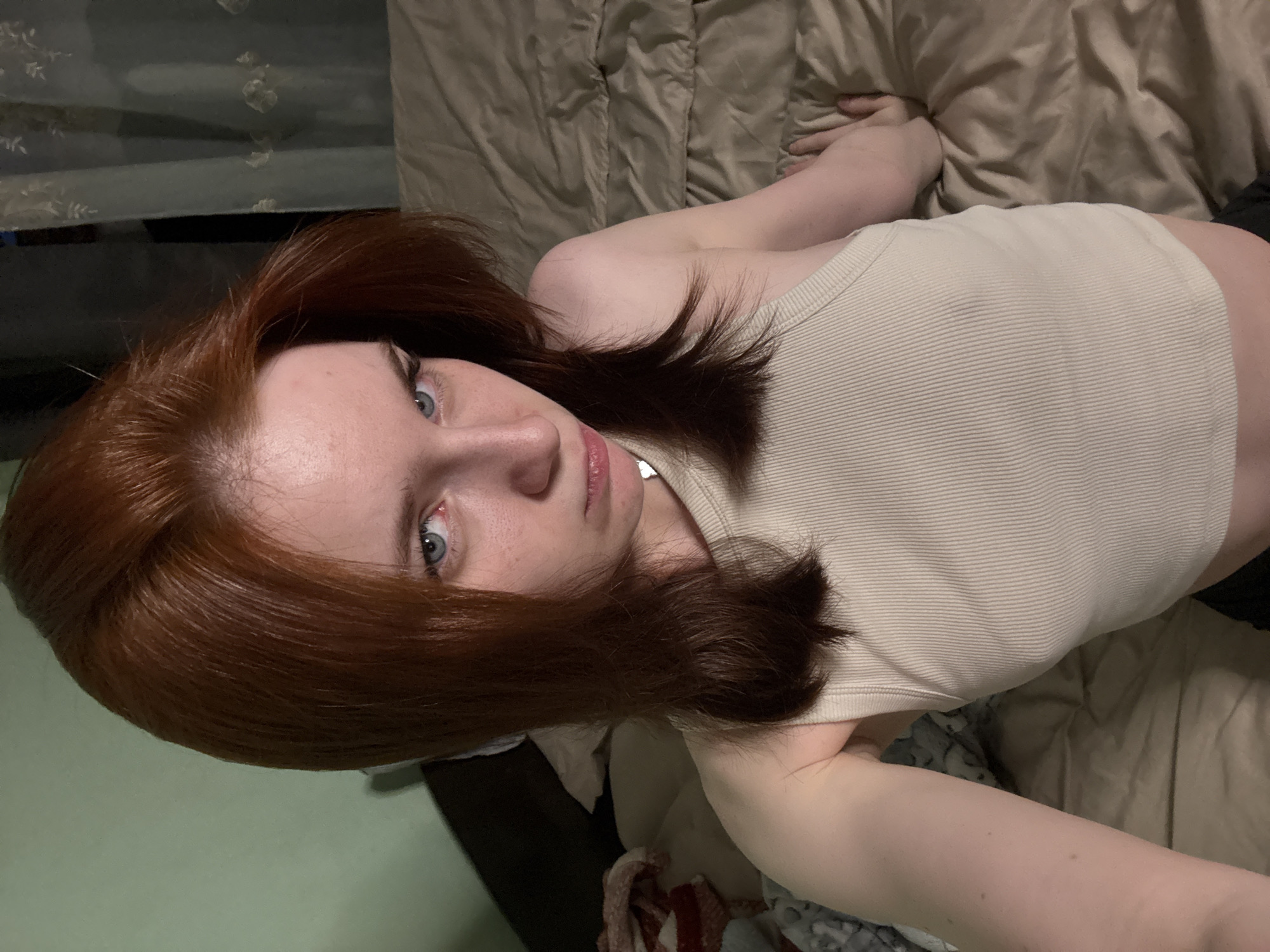 A red-haired person with pale skin and blue eyes is lying on a tan bed. They are wearing a beige ribbed tank top. The background includes bedding in varying shades of tan and gray. A small portion of a green wall is visible on the left.