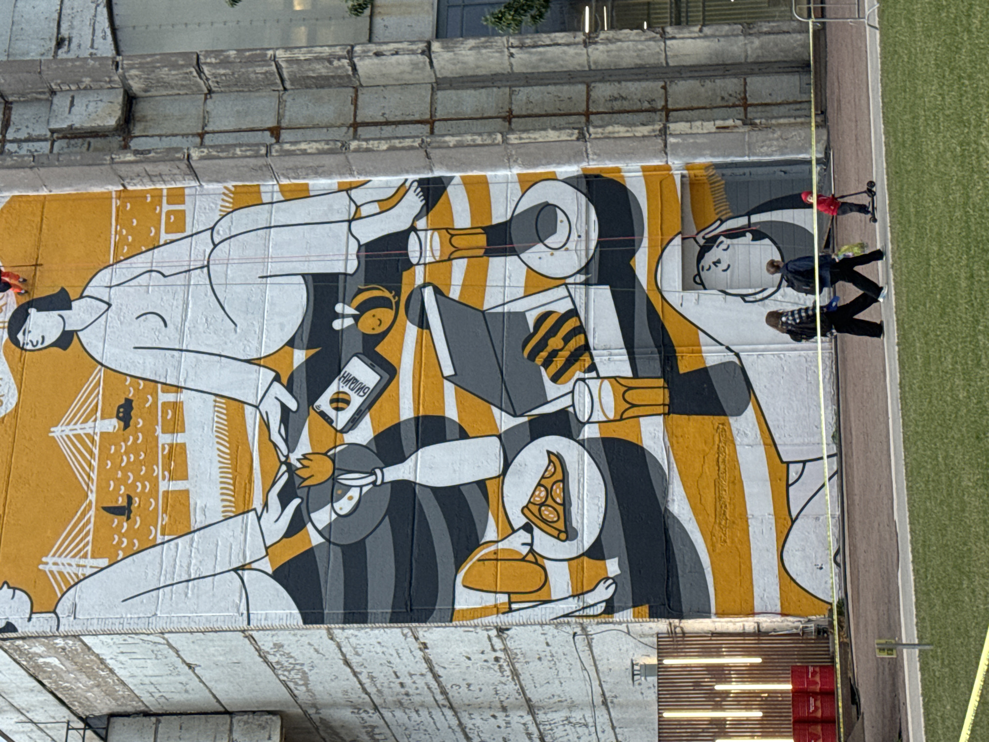 The image shows a large mural on the side of a building in Saint Petersburg, Russia. The mural is abstract and depicts a picnic scene with stylized figures, food, and a bee. In the foreground, there are three people walking: two adults and one child on a scooter. The building itself appears to be made of concrete and is weathered, suggesting an industrial or older urban environment. The area around the building is paved and has grass, indicating a public space. Yellow caution tape is visible across the foreground, separating the viewer from the area near the building.