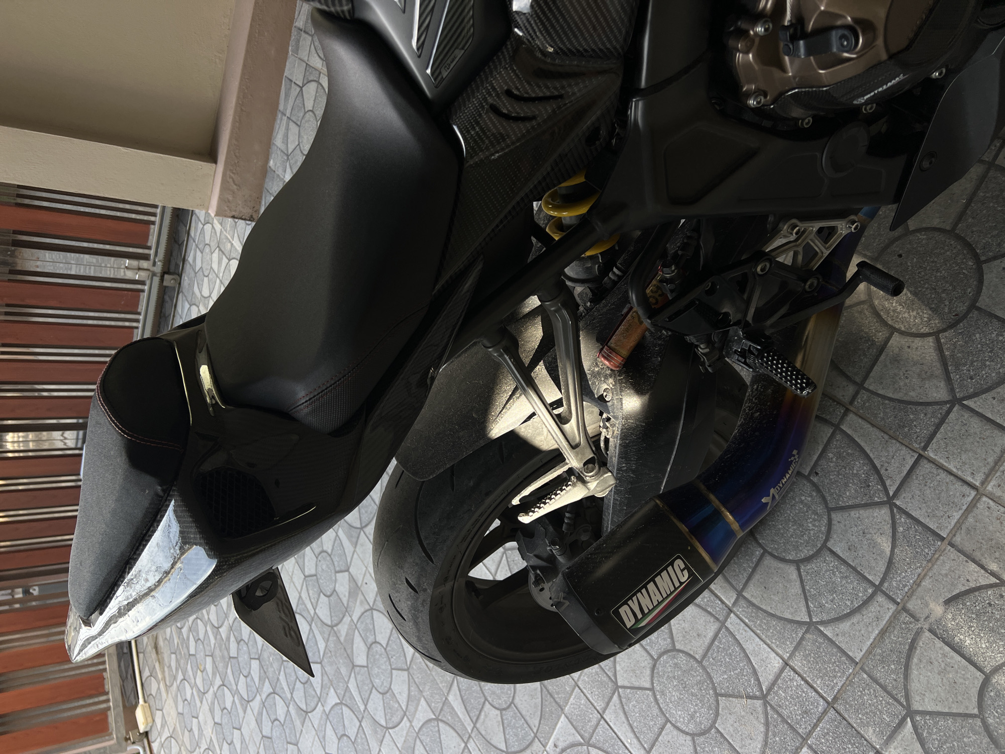 The image shows a close-up, angled view of the rear section of a black motorcycle, parked on a tiled patio. The motorcycle is the primary subject, with its dark seat, carbon fiber detailing, rear tire, and exhaust system prominently featured. The exhaust pipe has a heat-blued finish and bears the "DYNAMIC" logo in white lettering, along with a smaller "AKRAPOVIČ" logo.
The setting appears to be outdoors, with a tiled floor that has a circular pattern. A portion of a building or wall with horizontal blinds is visible to the left, suggesting it might be a residential or commercial outdoor space. The lighting indicates it is daytime, with sunlight casting shadows on the motorcycle and the ground.
There are no people visible in the image. The scene is static, with the motorcycle stationary. Notable details include the texture of the carbon fiber parts, the yellow suspension spring, and the wear on the tire. The overall impression is one of a well-maintained, sporty motorcycle. The text visible is "DYNAMIC" and "AKRAPOVIČ" on the exhaust.