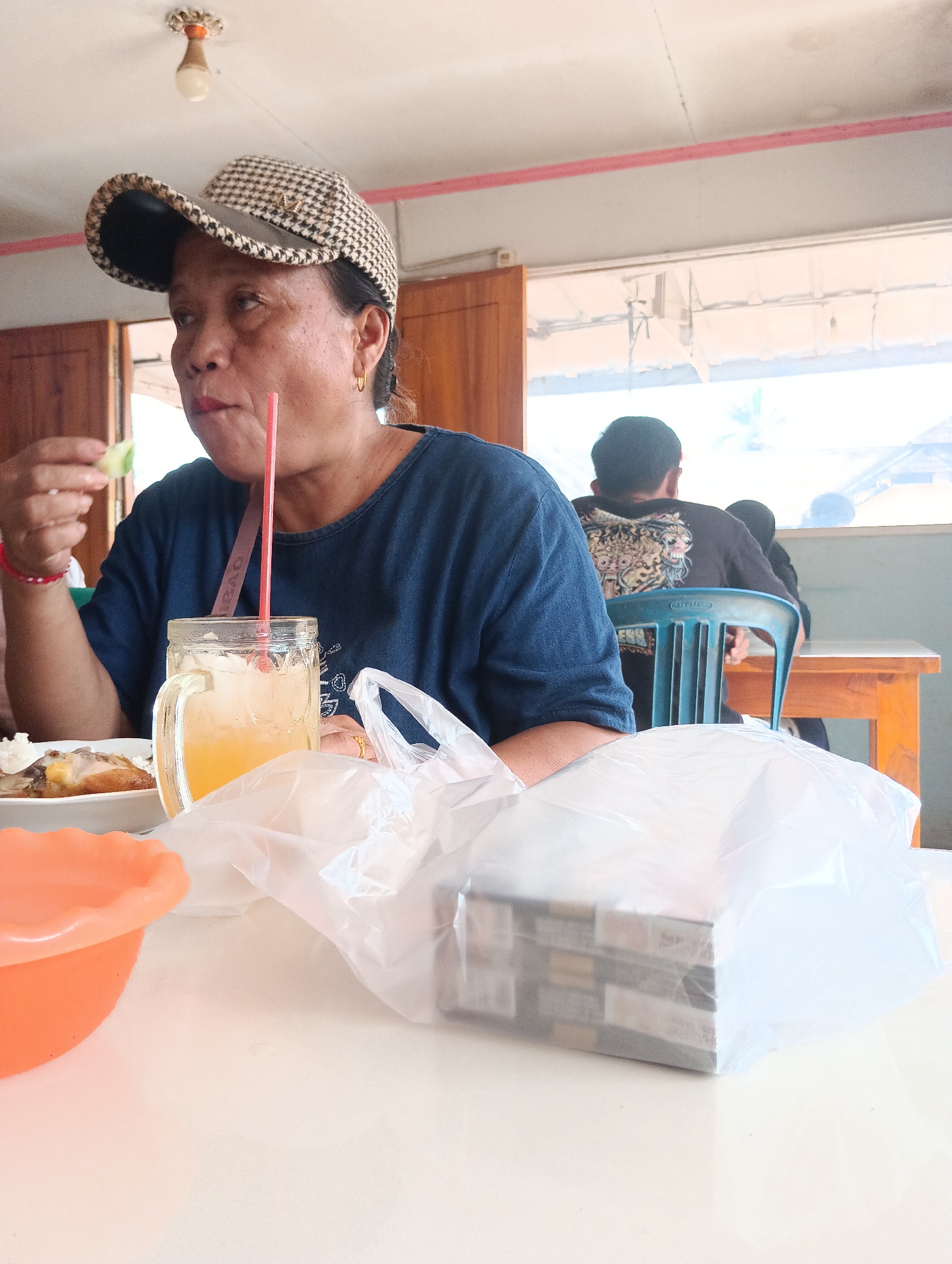 A medium shot captures a woman wearing a plaid cap and a dark blue shirt, seated at a white table in what appears to be an outdoor eatery in Binakarsa, Indonesia. She is holding a piece of cucumber in her left hand and has a straw in her drink. To her right, a glass jar filled with a yellow beverage and ice sits on the table, alongside a plastic bag containing what looks like stacked food items. In the background, two men are visible; one is seated at a blue chair facing away from the camera, while the other is partially visible to his right. The environment is simple, with wooden doors and window frames suggesting an open-air or semi-open space, and a bright ceiling light fixture is mounted overhead.