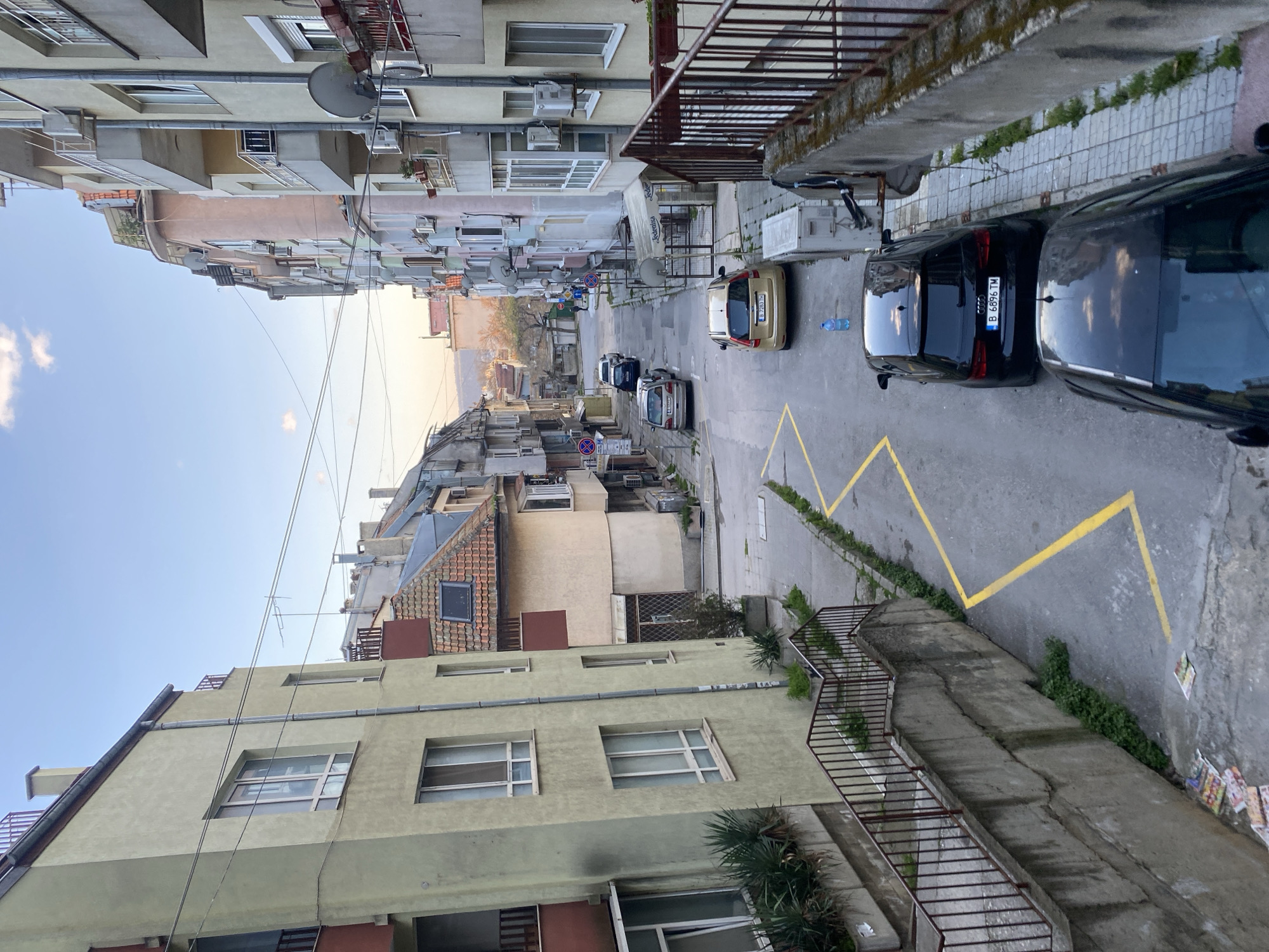 An elevated, slightly oblique view of a narrow street in Varna, Bulgaria. The image captures a street descending into the distance, flanked by closely situated apartment buildings. The architecture exhibits a mix of styles, with aged facades, protruding balconies, and satellite dishes adorning the buildings.
Cars are parked along the right side of the street, with an Audi bearing the license plate "B 6896 TM" visible. Yellow zig-zag lines mark the curb, indicating parking restrictions. Sidewalks are narrow, and concrete structures with metal railings border the right side.
The environment appears urban and densely populated, with visible signs of wear and aging. The light suggests either early morning or late afternoon. No individuals are immediately visible in the frame.