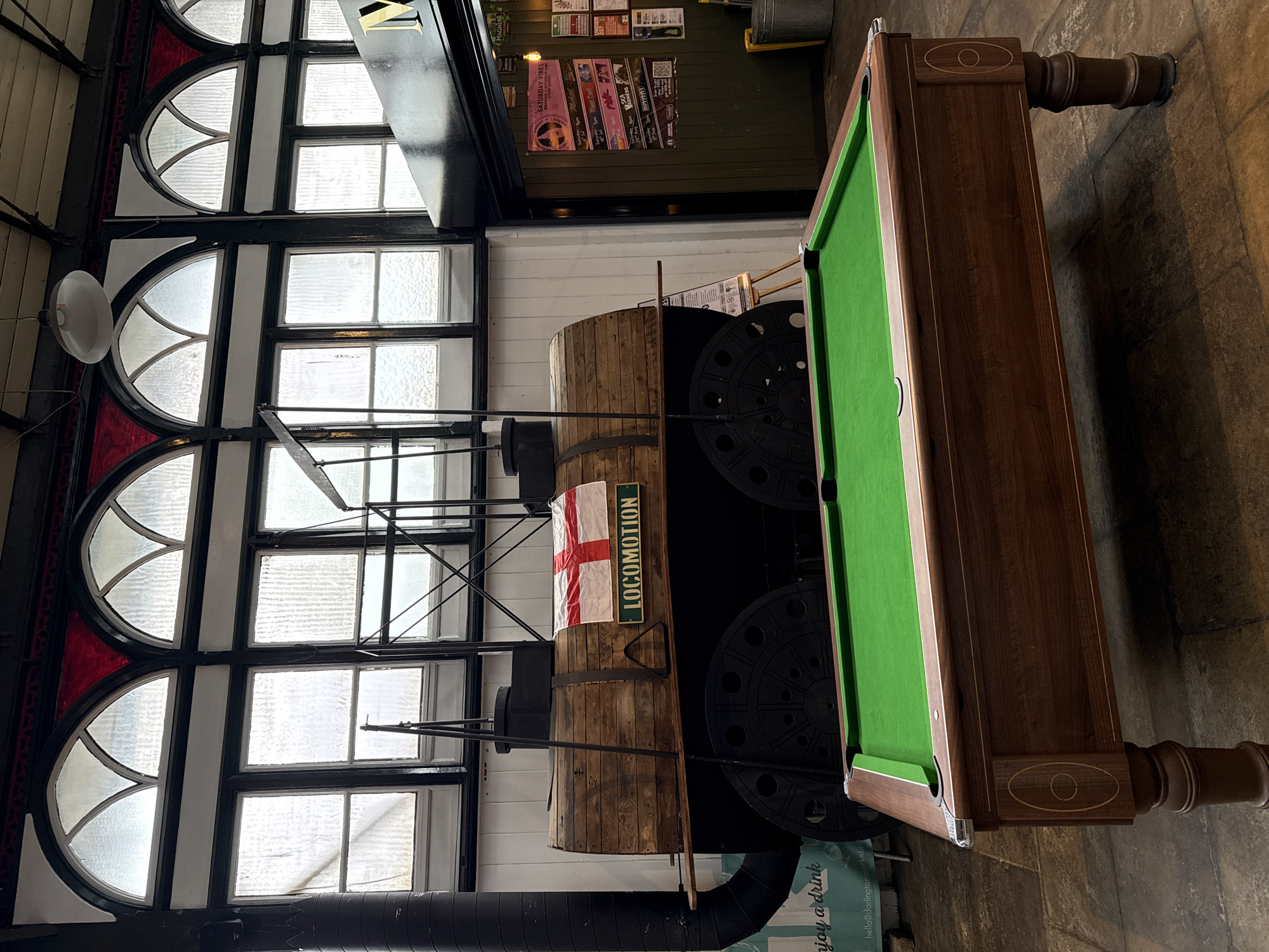 This image depicts an indoor scene, likely a pub or entertainment venue, featuring a pool table in the foreground and a decorative replica of an old steam locomotive in the background. The pool table is set up on a stone floor and is covered with green felt. It has a wooden frame and sturdy legs. Behind the pool table, a large, wooden structure resembling a steam engine boiler with the word "LOCOMOTION" and an English flag is displayed. The background also shows large, arched windows with glass panes, a dark ceiling with industrial lighting, and a wooden wall with posters and signage. The overall impression is a space that combines leisure and historical decor. This scene is located within the Darlington Market, a historic market hall in Darlington, United Kingdom.