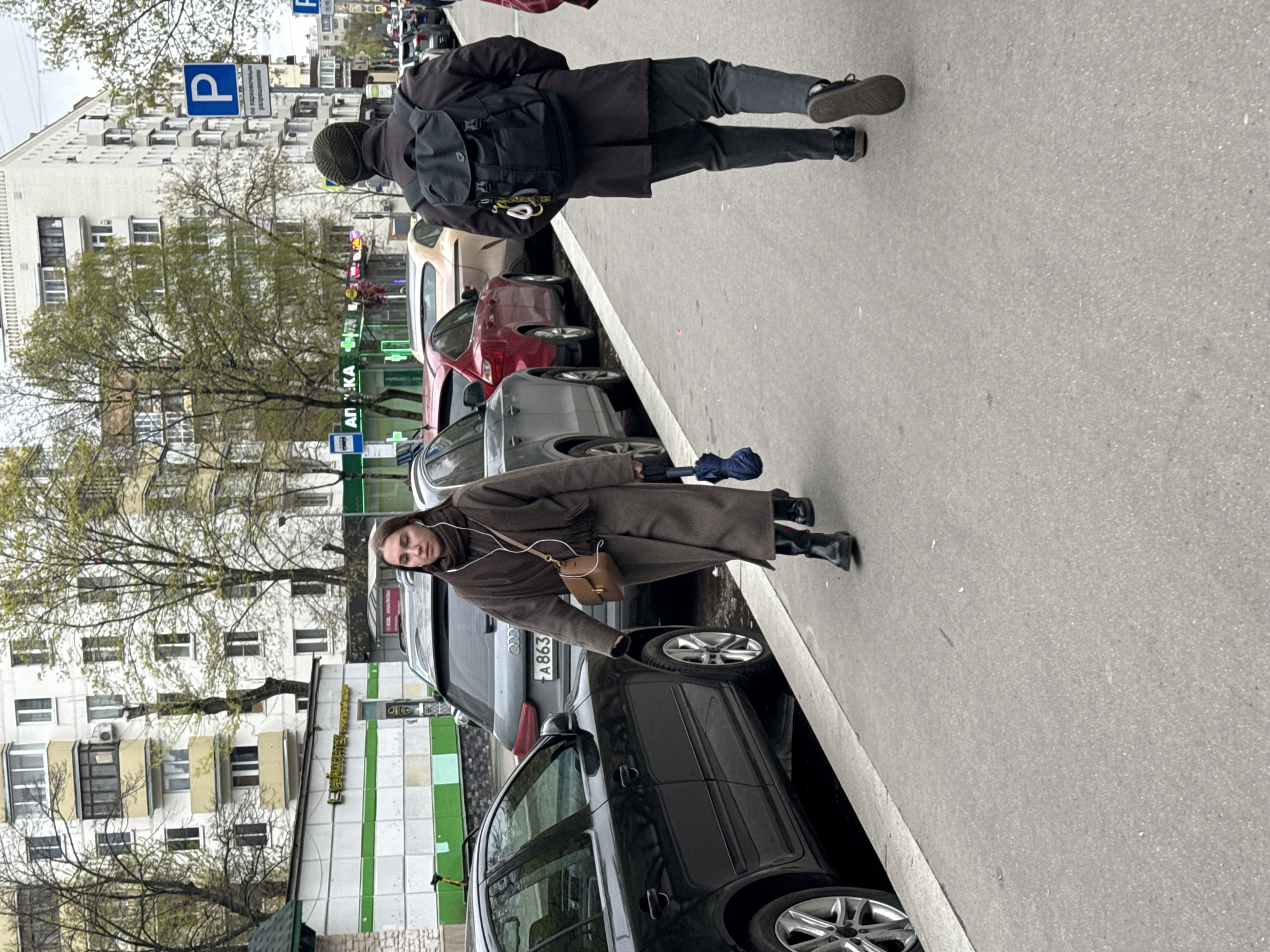 The image captures an overcast daytime street scene in what appears to be Moscow, Russia, given the Cyrillic text. A wide sidewalk runs parallel to a street lined with parked cars, with multi-story buildings forming the background.
Two individuals are present on the sidewalk. In the foreground, a woman stands facing the viewer, looking towards the left side of the frame. She wears a long, dark brown coat, black boots, and has wired earbuds in. She carries a small, light brown cross-body handbag and holds a closed blue umbrella in her left hand. Her expression is neutral. Further down the sidewalk, another person is walking away from the viewer, seen from behind. This individual wears a dark coat, dark pants, black sneakers, and a dark beanie. They are carrying a black backpack, which has yellow and silver carabiners attached.
Numerous cars are parked along the street. A dark sedan is prominent in the foreground, with an Audi sedan ("A863") behind it. Other vehicles, including a red car and a grey hatchback, are also visible.
The buildings in the background are multi-story, featuring many windows and air conditioning units. Some trees with sparse green leaves, suggesting early spring, are interspersed along the street and in front of the buildings.
Visible text includes a blue "P" parking sign with Cyrillic text reading "по разрешенным разрешениям" (by authorized permits). A green storefront sign prominently displays "АПТЕКА" (PHARMACY). Another building has a sign that appears to say "ЛЕЙКОВЫЙ" (LEYKOVYI), likely part of a business name. A blue street sign with a bus icon and Cyrillic text is also visible, possibly indicating a bus stop. The overall atmosphere is that of a typical urban day.