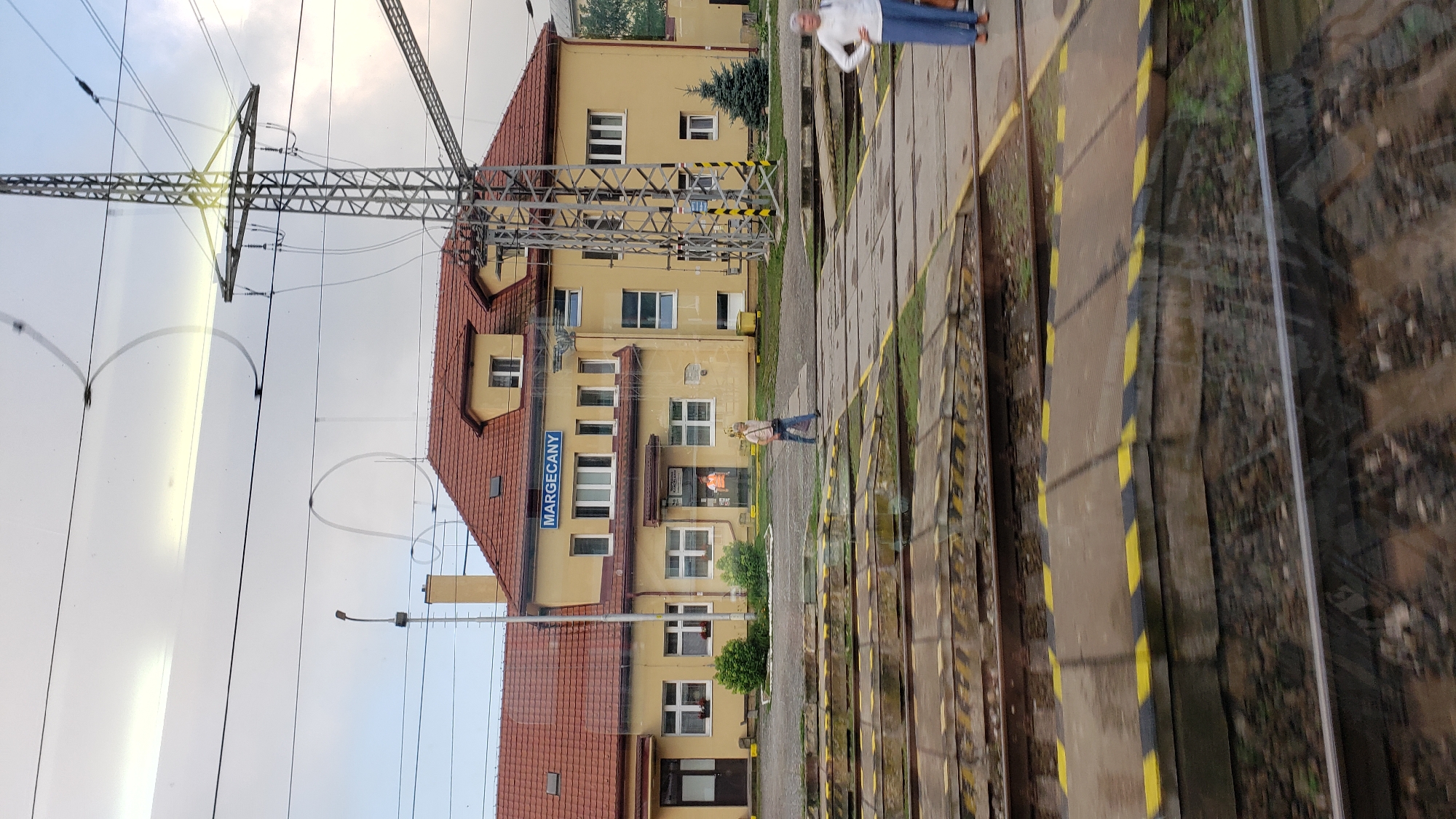 The image shows the Margecany train station in Margecany, Slovakia. The main building of the station is a two-story yellow structure with a red tiled roof. A blue sign above the entrance reads "MARGECANY". There are several windows on the facade, and a person wearing an orange vest is visible through a glass door. In front of the building, there is a paved area with train tracks and platforms. Two women are standing on the platform, and one woman is walking across the tracks. A tall metal electricity pylon stands to the right of the station building. The sky is overcast.