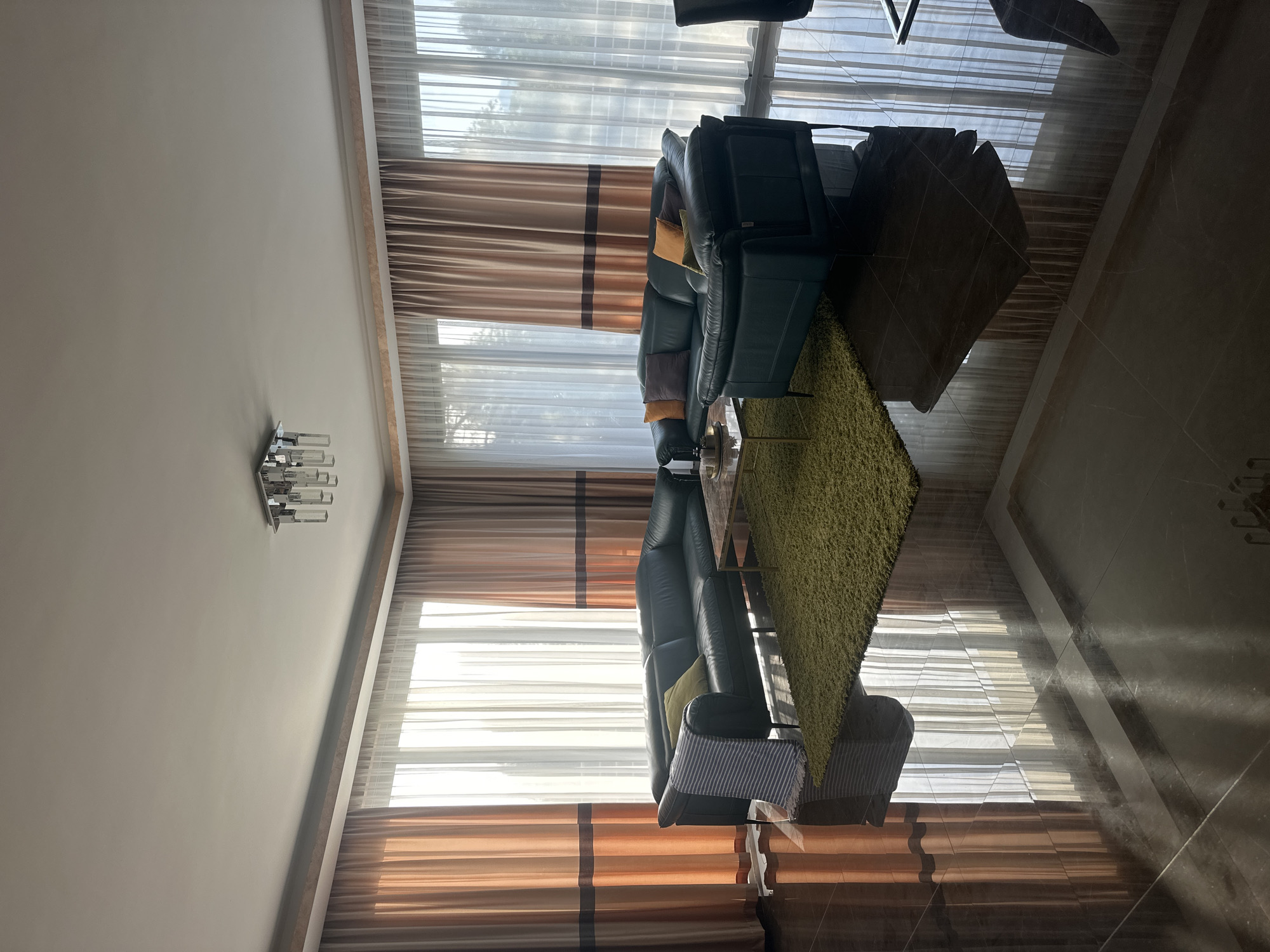 A well-lit, modern living room in Brca, Montenegro features a large, dark green leather sectional sofa arranged in an L-shape around a glass-topped coffee table. A vibrant green shag rug sits in the center of the seating area. The room is illuminated by natural light streaming through large windows covered by sheer white curtains and accented with floor-to-ceiling curtains in shades of peach and brown. A contemporary silver chandelier hangs from the ceiling. The room's floor is made of highly polished, dark marble tiles that reflect the light and the furnishings. On the sofa, several throw pillows in various colors, including yellow, orange, and brown, are visible, along with a striped throw blanket draped over one section of the sofa. The environment appears clean and organized, with no people present.