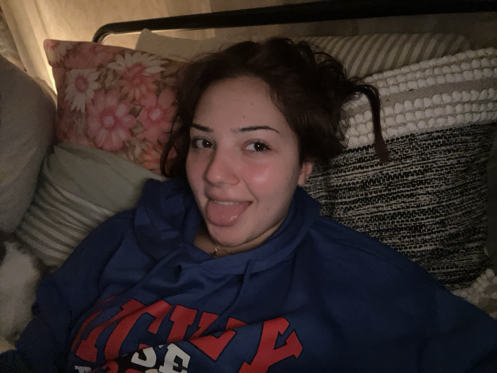 A young woman lies in bed surrounded by pillows. She wears a blue hooded sweatshirt with red lettering. The woman is sticking her tongue out. Her dark hair is pulled up messily. Pillows behind her include a floral print, a striped pattern, and a woven design. The lighting is dim.
