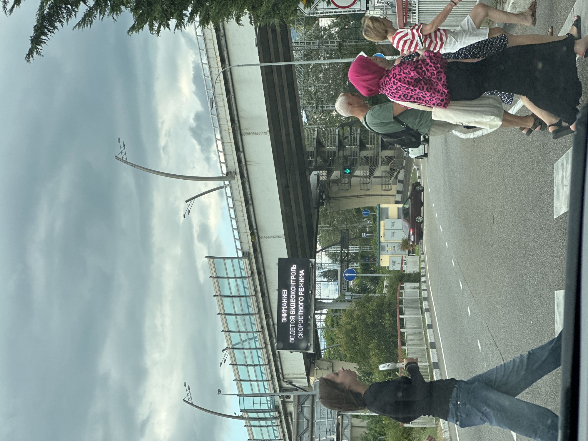 The image captures a pedestrian crossing on an overcast day in Sochi, Russia, likely near the Olympic Park area given the architectural style of the overpass and the presence of a large digital billboard. The billboard, positioned under a multi-lane overpass, displays a message in Russian: "ВНИМАНИЕ! ВЕДЕТСЯ ВИДЕОКОНТРОЛЬ СКОРОСТНОГО РЕЖИМА" which translates to "ATTENTION! VIDEO CONTROL OF THE SPEED REGIME IS BEING CONDUCTED."
Several people are visible crossing the street. In the foreground left, a young woman with dark hair walks from left to right, holding a portable fan. To her right, a group of three individuals are crossing from right to left. The first person in this group is a man with grey hair wearing a green t-shirt and a backpack. Next to him is a woman wearing a bright pink headscarf and a leopard-print top, with a long black skirt. Behind her, a woman with blonde hair wears a striped t-shirt and white shorts. A maroon car is visible in the background on the road. The environment includes a paved road with white lane markings, a sidewalk with a metal railing, and dense green foliage bordering the road and beneath the overpass. The sky is heavily clouded, contributing to the dim lighting.