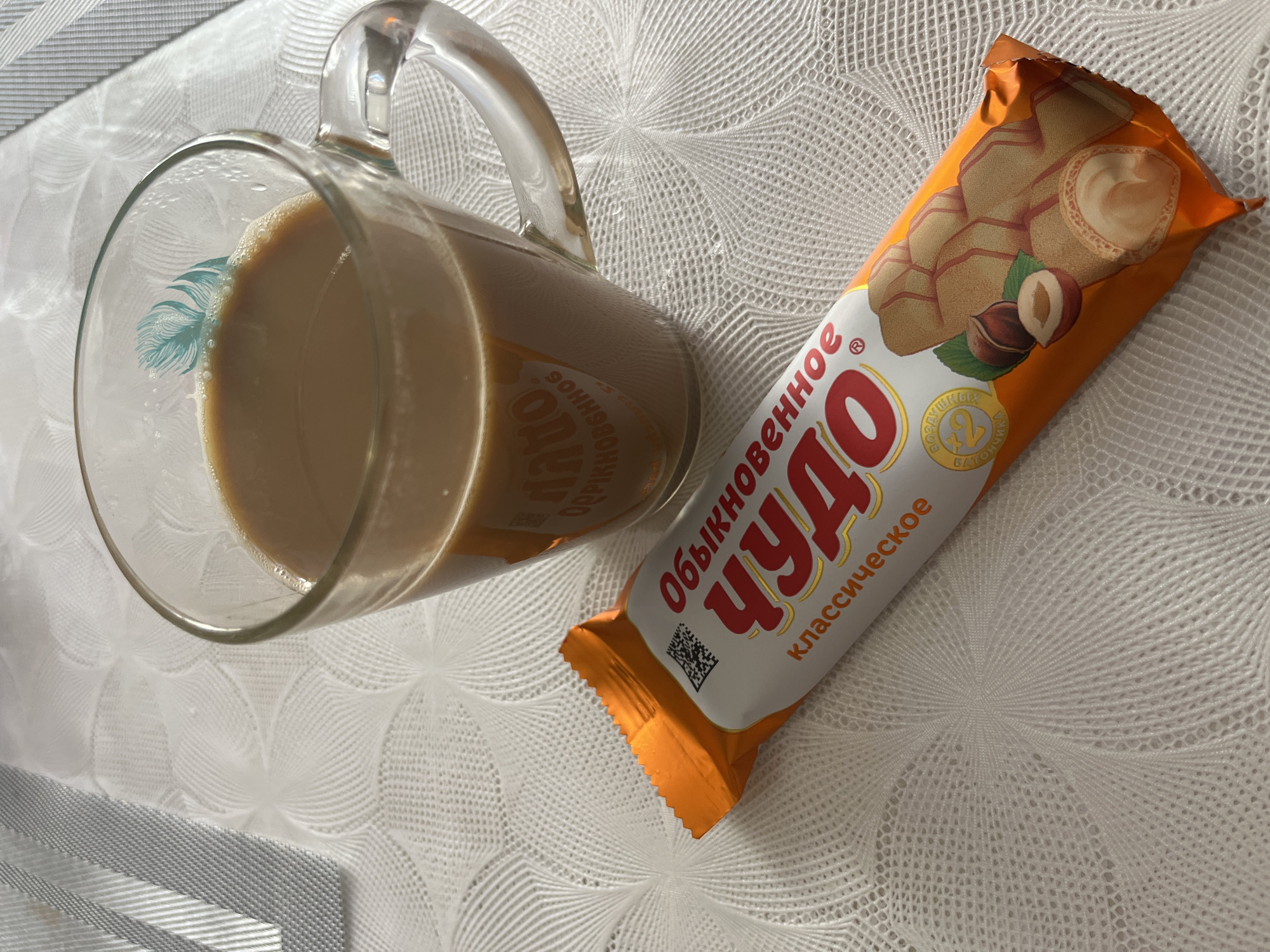 A clear glass mug filled with a light brown beverage rests on a white, patterned surface. Next to the mug is a package of "Chudo" brand wafer snacks. The snacks are in an orange and white wrapper. The text on the package is in Russian. The image was likely taken in Kaspiysk, Russia, as the product is Russian. The mug and package are on a white surface that may be a tablecloth. No people are visible.