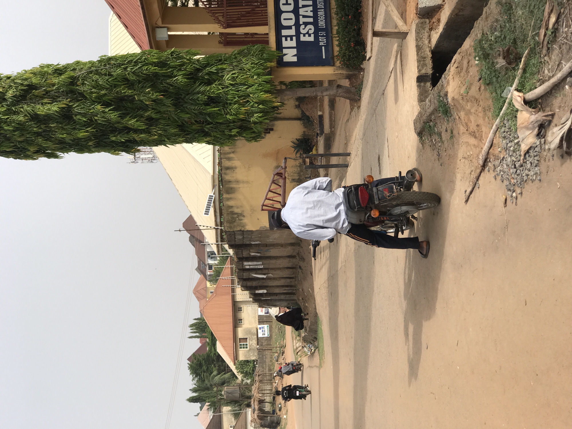 This daytime image depicts a street scene in Abuja, Nigeria, specifically in the Lokogoma District, as confirmed by a visible sign. The environment appears to be a mix of residential and developing areas.
In the foreground, a person is seated on a motorcycle, facing away from the viewer. The individual wears a light-colored, long-sleeved shirt and dark trousers with a prominent orange stripe. The motorcycle is dark-colored, equipped with red taillights and amber turn signals. The road directly beneath the motorcycle is an unpaved, light brown dirt surface.
To the left of the road, a series of buildings line the street. A notable structure features a light yellow facade and a red-tiled roof. Standing tall next to this building is a very slender tree with dense, vibrant green foliage. Attached to a pole near the tree is a dark blue sign with white lettering. The text clearly reads: "NELOC ESTATE" at the top, and "PLOT 51 LOKOGOMA DISTRICT" underneath, providing a precise location cue.
To the right of the motorcycle and the road, the terrain is rough, featuring a raised dirt embankment with sparse green vegetation and scattered gravel. A long, thin piece of wood or branch lies on the ground, and a visible concrete-lined drainage channel runs along this side.
In the mid-ground and background, the street extends, revealing more residential buildings with various roof types, including red-tiled and flat designs. Several other motorcycles are visible further down the road, indicating movement and activity. The sky above is bright but appears hazy or lightly overcast, casting soft shadows. The scene portrays a functional street within a residential area.