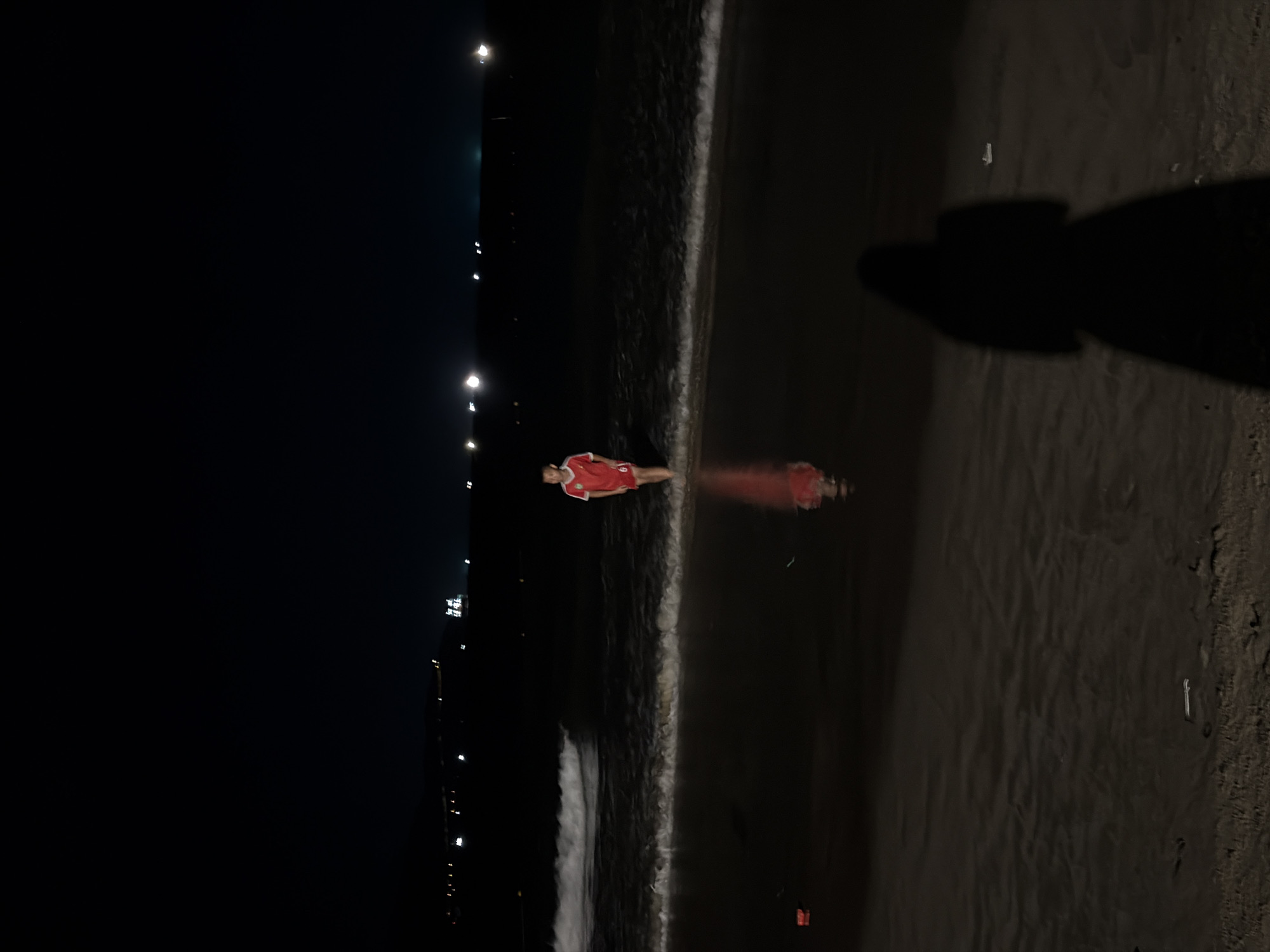 At night on a beach in Da Nang, Vietnam, a young boy wearing a red and white soccer jersey stands in the shallow ocean water. The waves are breaking gently around his legs. His reflection is visible in the wet sand. In the foreground, a shadow of a person, likely the photographer, is cast on the sand. In the background, distant lights from the city illuminate the dark sky.