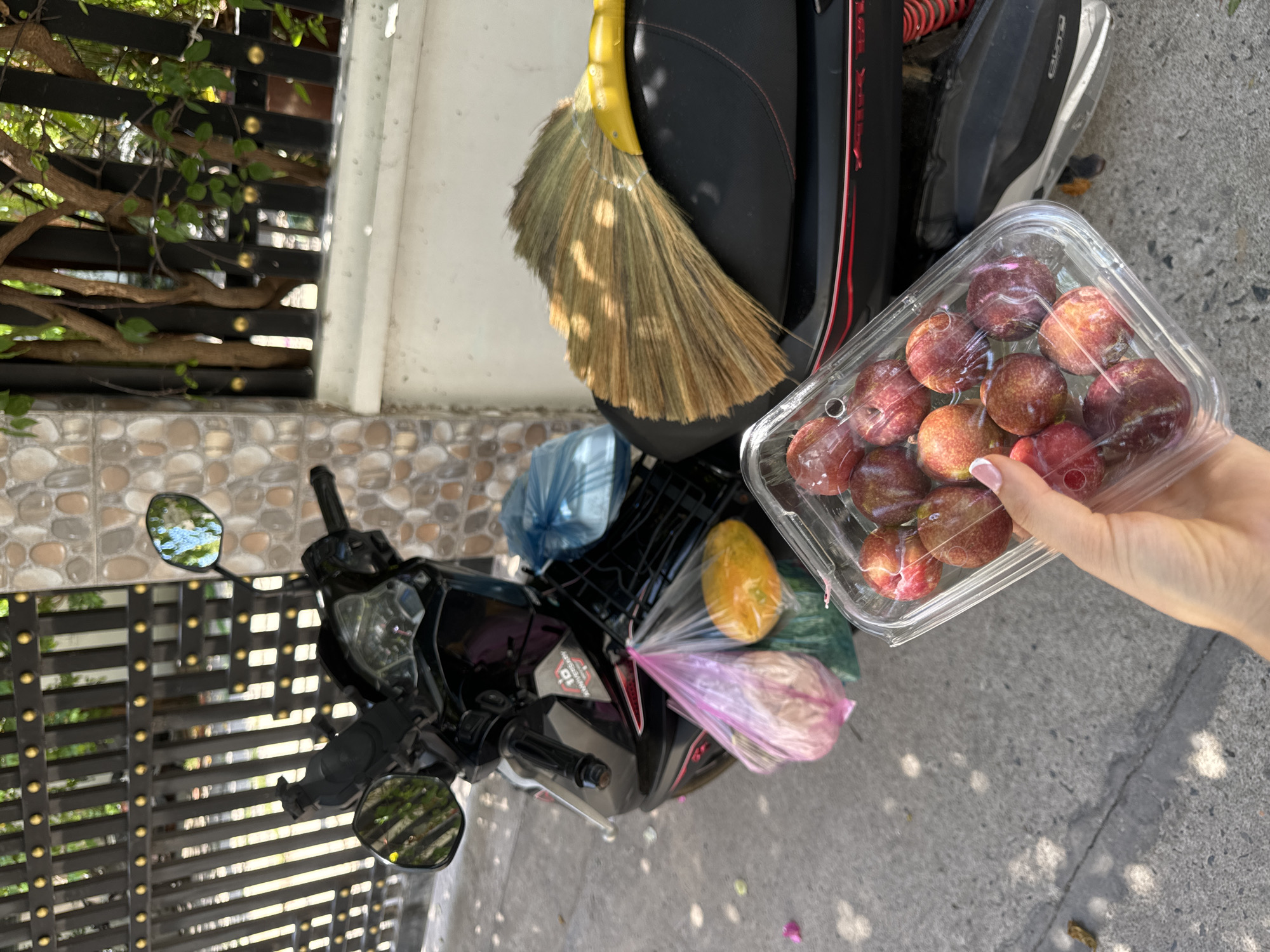 A street scene in Mũi Né, Vietnam, captures a black scooter parked on a sidewalk with a brown broom attached to the seat. Various items, including fruits in plastic bags and an open transparent container holding approximately 20 plums, are secured to the scooter. A person's right hand is visible, holding the container of plums. In the background, a patterned wall and foliage are visible behind a metal gate. The sidewalk shows shadows and debris.