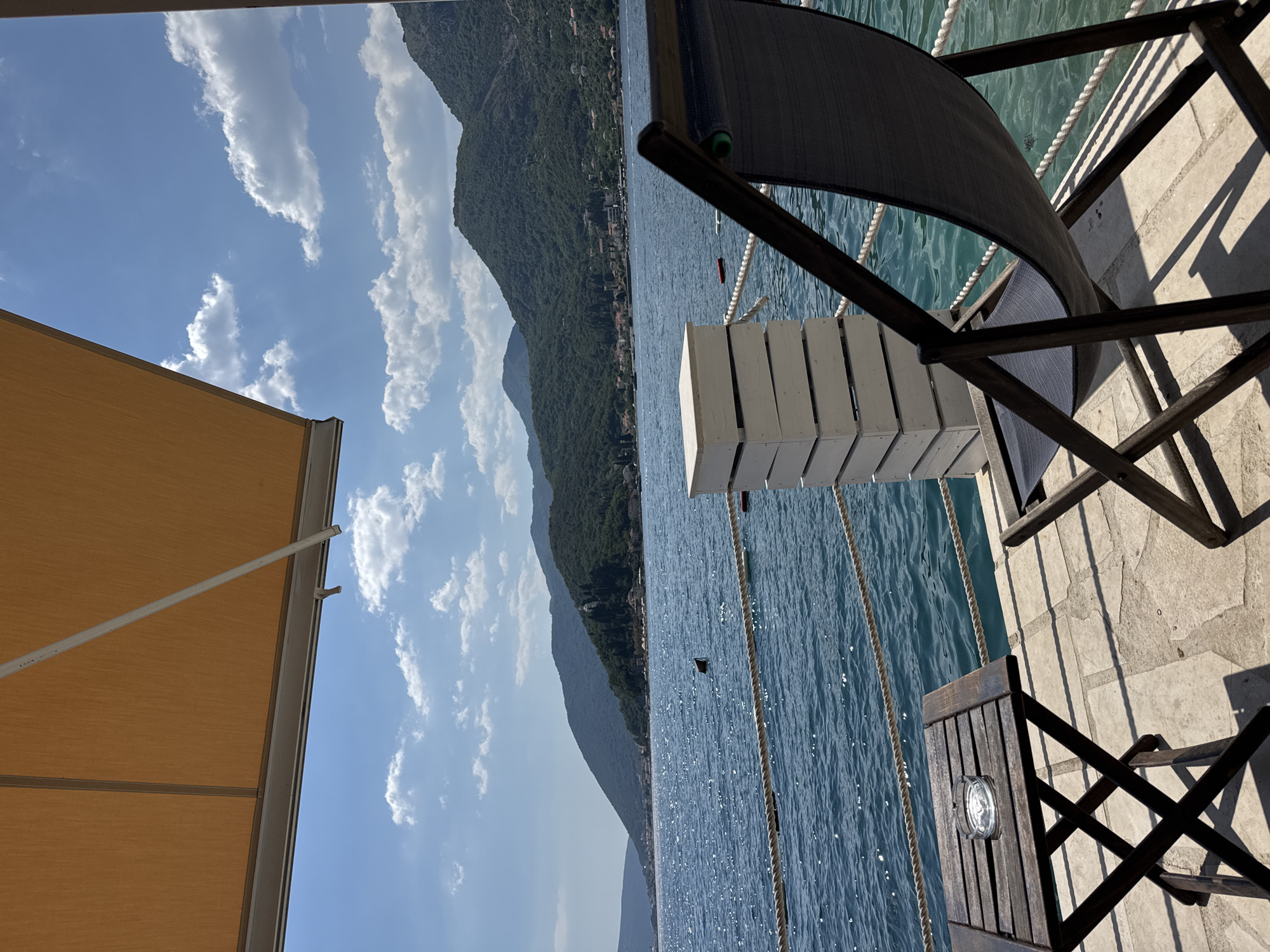 The image depicts a serene outdoor seating area with a view of a body of water and distant hills. In the foreground, a dark wooden folding table with an ashtray sits on a stone-tiled patio. Next to it is a dark-colored lounge chair, partially obscuring the view. A white wooden railing with a thick rope cordons off the seating area from the water. Beyond the railing, the bright blue water is dotted with white reflections from the sun. In the middle ground, a town with red-tiled roofs is nestled along the shore, backed by lush green hills that rise into the distance under a bright blue sky with scattered white clouds. The scene suggests a tranquil vacation spot.