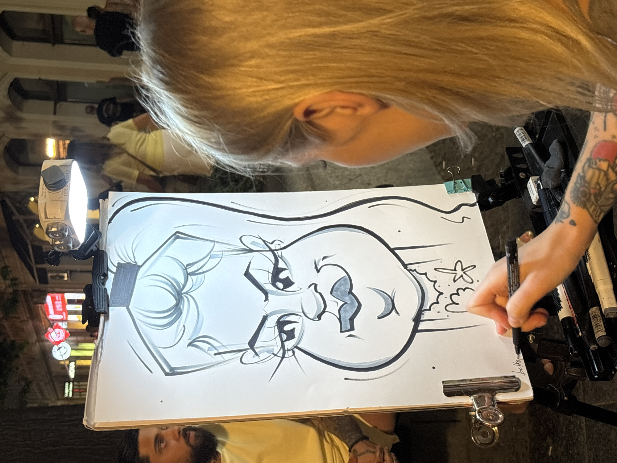 The image shows a street artist in Belgrade, Serbia, drawing a caricature of a man. The artist, a woman with blonde hair, is in the foreground, her back to the camera, as she sketches on a large white board propped up on an easel. She is holding a black marker in her right hand and appears to be adding finishing touches to the drawing. The caricature itself is a stylized portrait of a man with exaggerated features, including a large head, prominent eyes, and full lips. A light source, possibly a portable lamp, is mounted above the drawing board, illuminating the artwork. In the background, the artist's subject, a man with dark hair and a beard, is visible to the left, looking towards the drawing. He is wearing a yellow t-shirt. Behind them, a bustling street scene unfolds with blurred figures of people walking and illuminated shop signs, suggesting the setting is likely a pedestrian area or a lively street in the city at night. The general ambiance is that of an outdoor urban environment.
