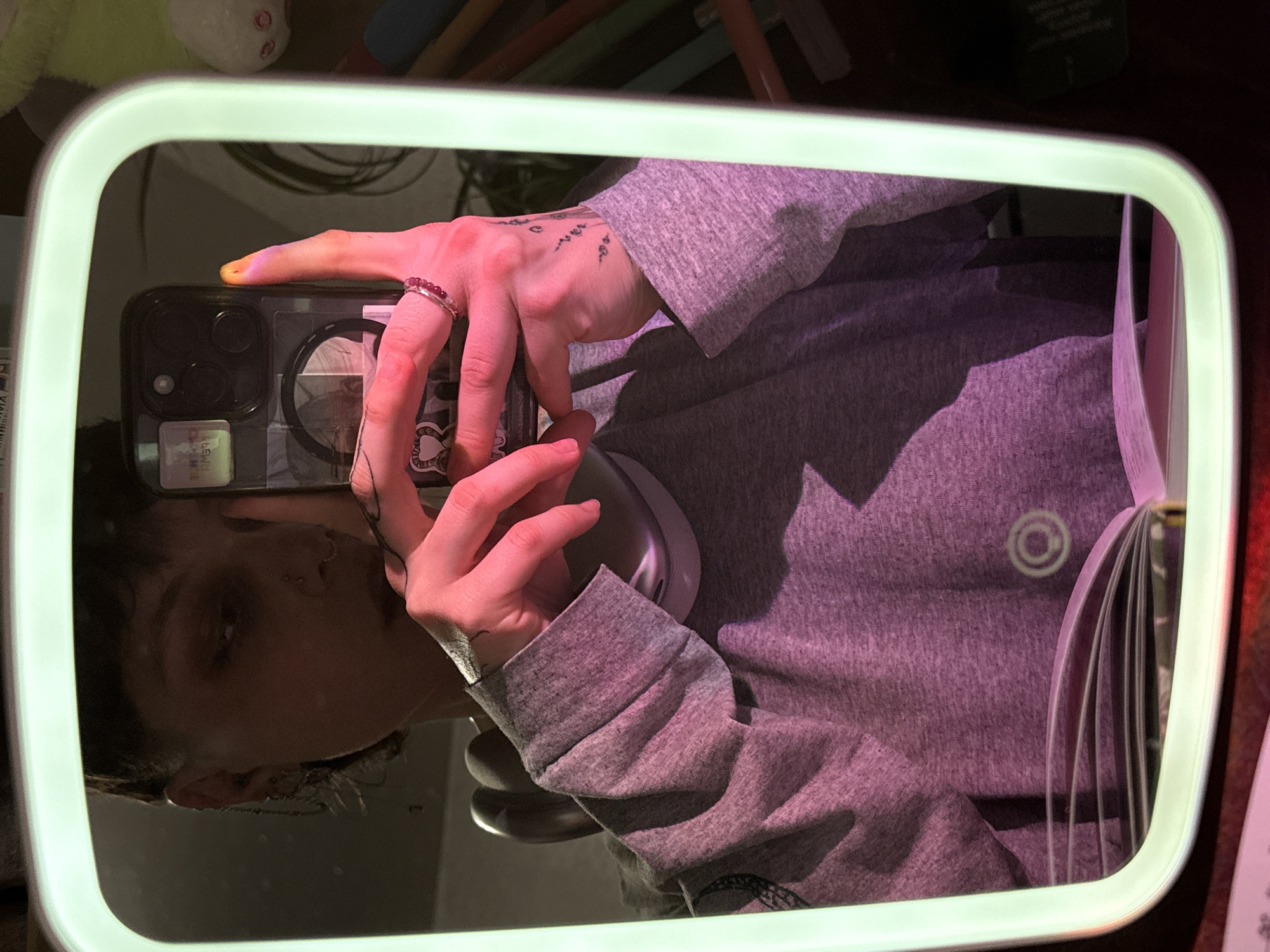 A person wearing a gray sweater takes a selfie in a rectangular mirror with a glowing green frame. The reflection shows the person's face partially, their hand holding a smartphone, and part of their torso. The person has tattoos on their hand and is wearing a ring on one finger. The smartphone has a decorative case. Behind the mirror are various objects, including a notebook and writing utensils. The photograph was taken indoors, presumably in Russia. The exact location in Saint Petersburg is impossible to determine based on the image.