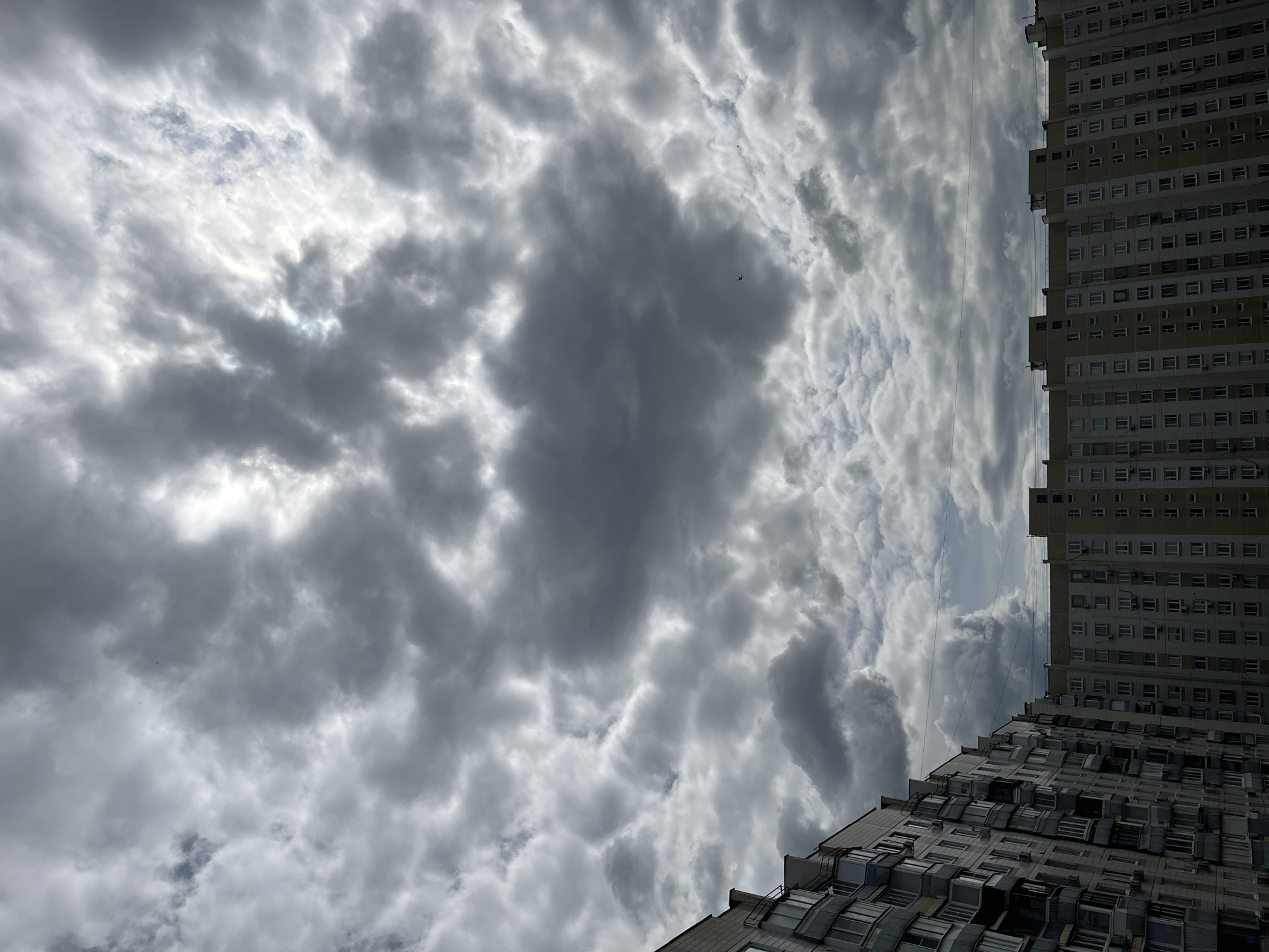 The image depicts a view of apartment buildings under a cloudy sky. The buildings are tall, multi-story structures with numerous windows and balconies, suggesting an urban residential area. The sky is filled with dense, gray clouds, with some patches of brighter light breaking through, indicating possibly overcast or stormy weather. There are no people or animals visible in the frame. The architecture of the buildings, with their repetitive window patterns and balconies, is characteristic of mass-produced Soviet-era or post-Soviet housing blocks. The scene appears to be captured from a slightly elevated perspective, possibly a window or another building. The image does not provide specific clues to pinpoint the exact location in Moscow, Russia.
