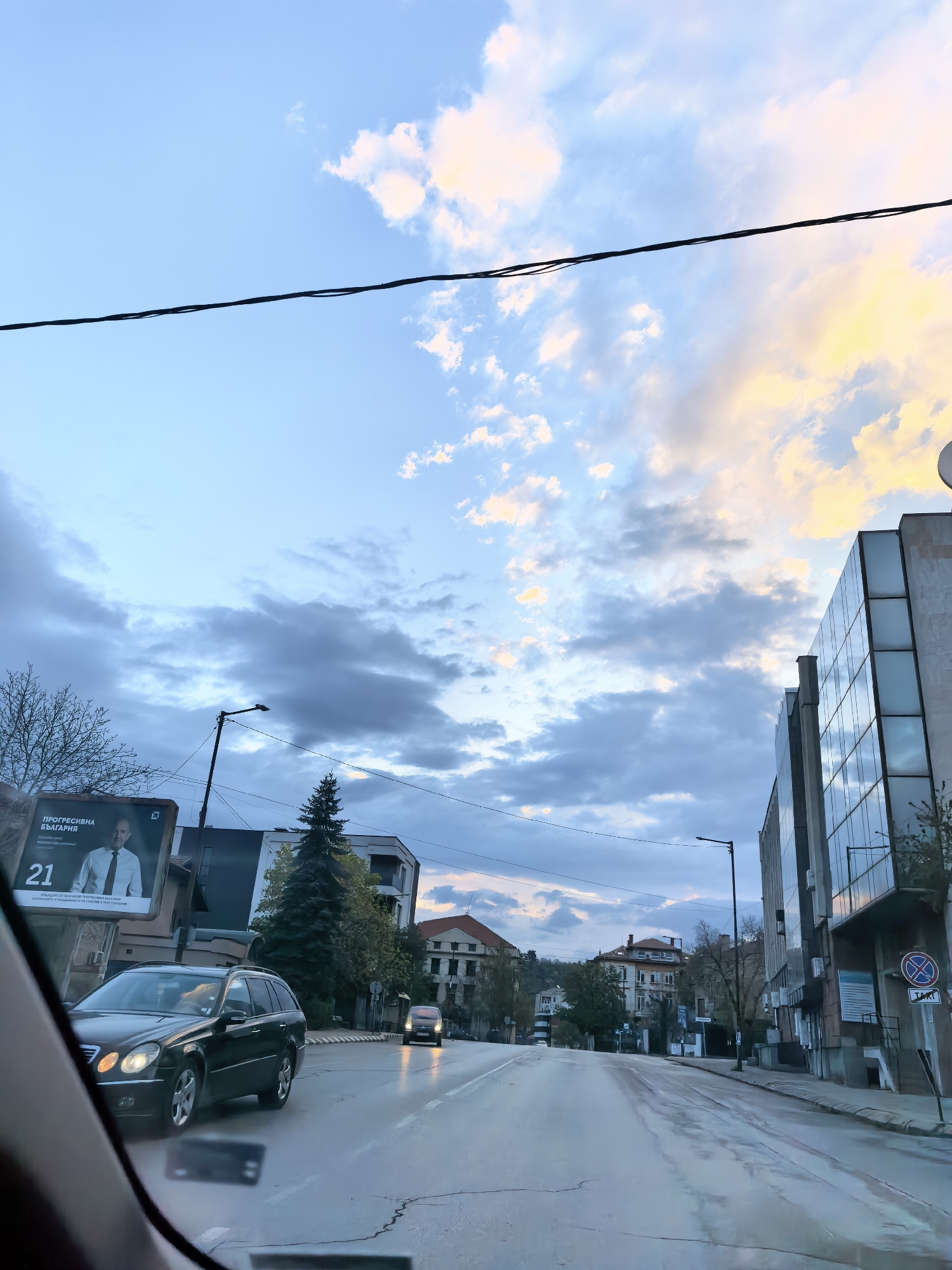 A street view from inside a car, showing wet asphalt reflecting the sky. On the left, a black station wagon passes a billboard with a man's face and the text "ПРОГРЕСИВНА БЪЛГАРИЯ" (Progressive Bulgaria). Straight ahead, the road leads between buildings, some with orange tile roofs. On the right, a tall building with mirrored glass panels is visible, along with a street sign indicating parking restrictions. The sky is cloudy with shades of blue and yellow. A power line crosses the upper part of the frame.