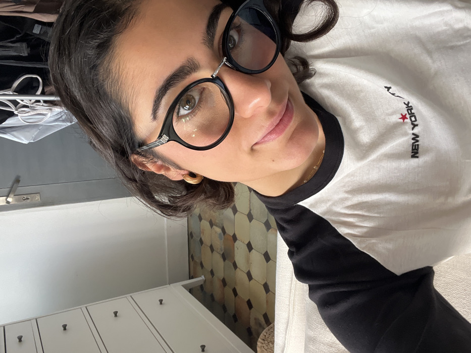 A young adult female is depicted in a medium close-up, centered slightly right, making direct eye contact with a subtle smile. She has dark, wavy hair and wears black-framed, round prescription glasses featuring a small silver detail on the upper left frame. Her left ear displays a gold-colored hoop earring, and a thin gold-colored necklace is visible around her neck. She is dressed in a white t-shirt with black raglan sleeves, embroidered with the text "NEW YORK" in black, followed by a red star.
The background establishes an interior residential setting, identifiable as an apartment in Paris, France, due to distinctive architectural elements. The floor features traditional hexagonal or octagonal terracotta "tomettes" tiles in a pattern of varying light brown, beige, and dark brown shades. To the left, a portion of a white chest of drawers with four visible black knobs is present. A segment of a white sofa armrest is visible in the lower left. Towards the center-right background, a grey-blue interior door is visible, fitted with a characteristic Parisian-style metallic lever handle. In the upper right quadrant, dark clothing items are partially visible, hanging on a rack or hangers.