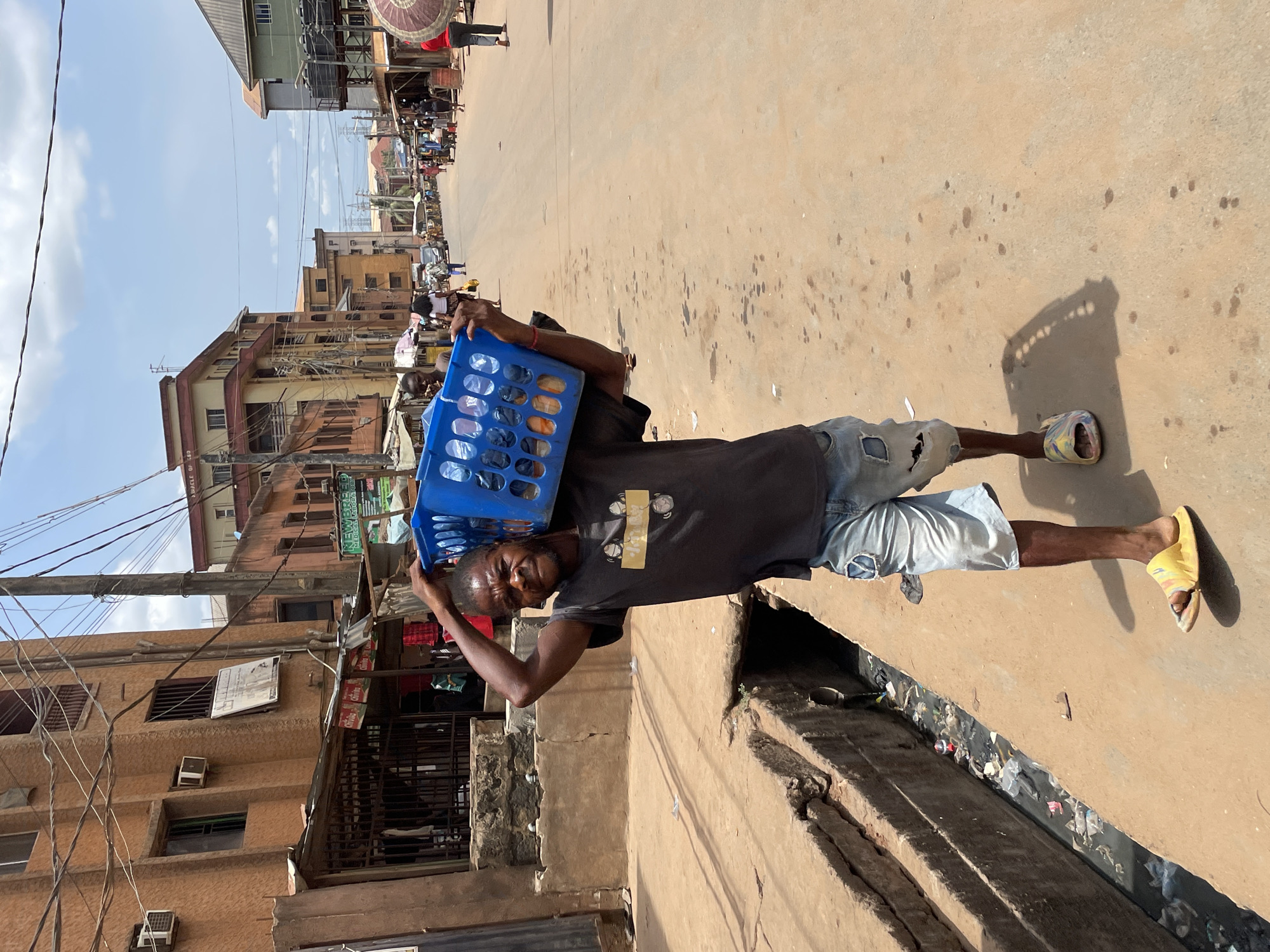 The image captures a vibrant street scene in Onitsha, Nigeria, under bright, clear daylight. The primary subject is a man of African descent, positioned towards the center-right of the frame, facing the viewer. He is carrying a blue plastic crate, filled with what appear to be numerous pairs of sandals or slippers, balanced on his head. He wears a dark t-shirt with a yellow graphic on the chest, ripped light-wash denim shorts, and yellow slide sandals. His expression is one of acknowledgement, possibly a slight smile, as he looks towards the camera.
The setting is a busy street, with a worn, dusty asphalt surface. To the man's left, a wide, open drainage ditch runs parallel to the road, filled with visible trash and debris. In the background, the street stretches further, with other people and activity suggesting a bustling market or commercial area. Multi-story buildings, varying in age and style, line the left side of the street. Numerous power lines and utility cables crisscross the sky above the thoroughfare.
Notable details include the strong shadows cast by the bright sun, indicating a mid-day time. The architecture and street vendors (implied by the man's activity) are characteristic of an urban Nigerian environment.
Visible text includes a sign on one of the buildings on the left that reads "NEW BLESSED MEDICAL STORE," followed by a phone number and "24H SERVICE," and "DRUGS." Additionally, several smaller signs feature the "Chivita" logo.